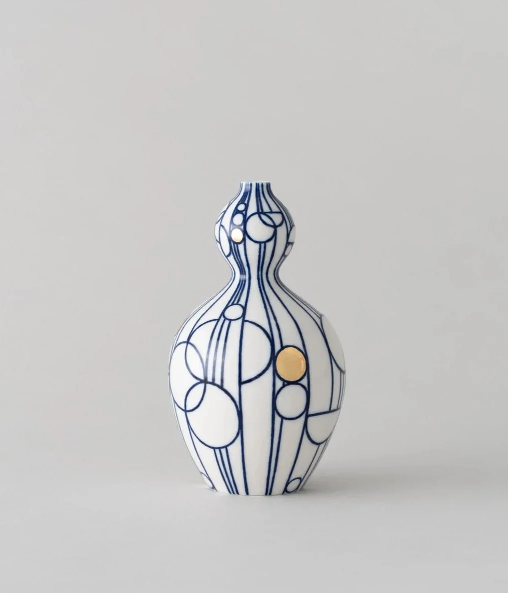 White ceramic vase with blue circular and line patterns, featuring a few gold accents, placed on a plain gray background.