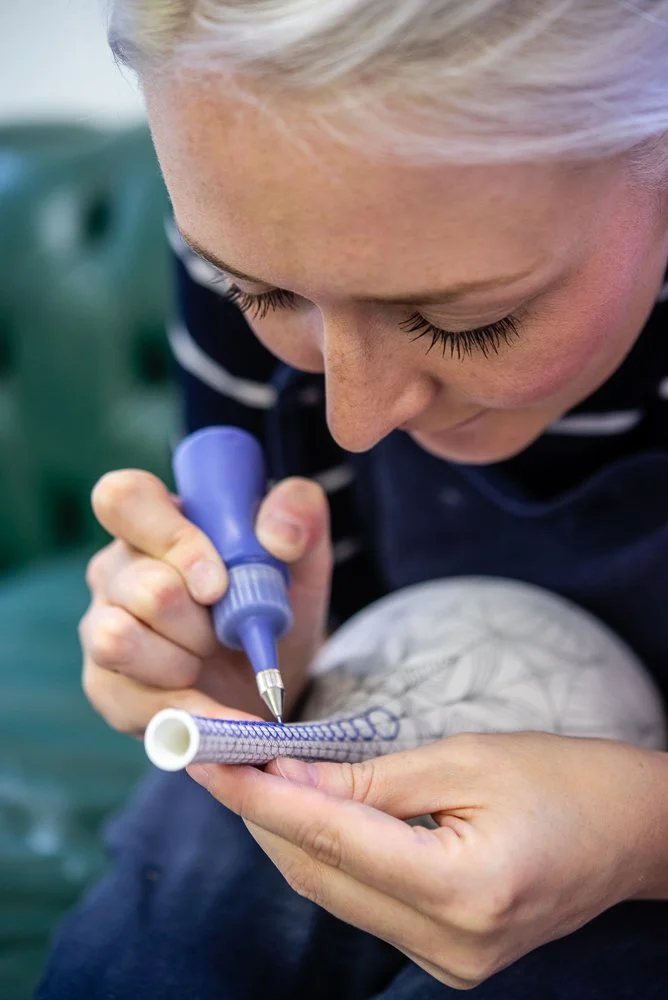 A woman is carefully designing on a white cylindrical object, possibly a ceramic or porcelain pipe, using a blue marker, with a close focus on her careful work.