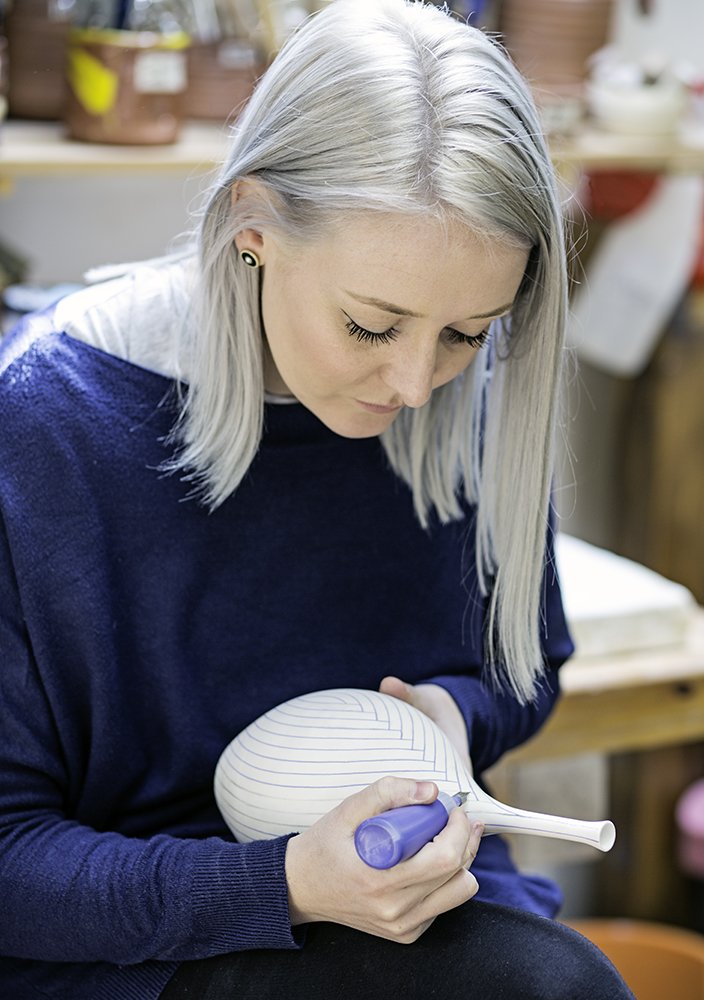 A woman with platinum blonde hair and earrings, focused on decorating a white ceramic vase with blue lines using a blue marker in a craft workshop.