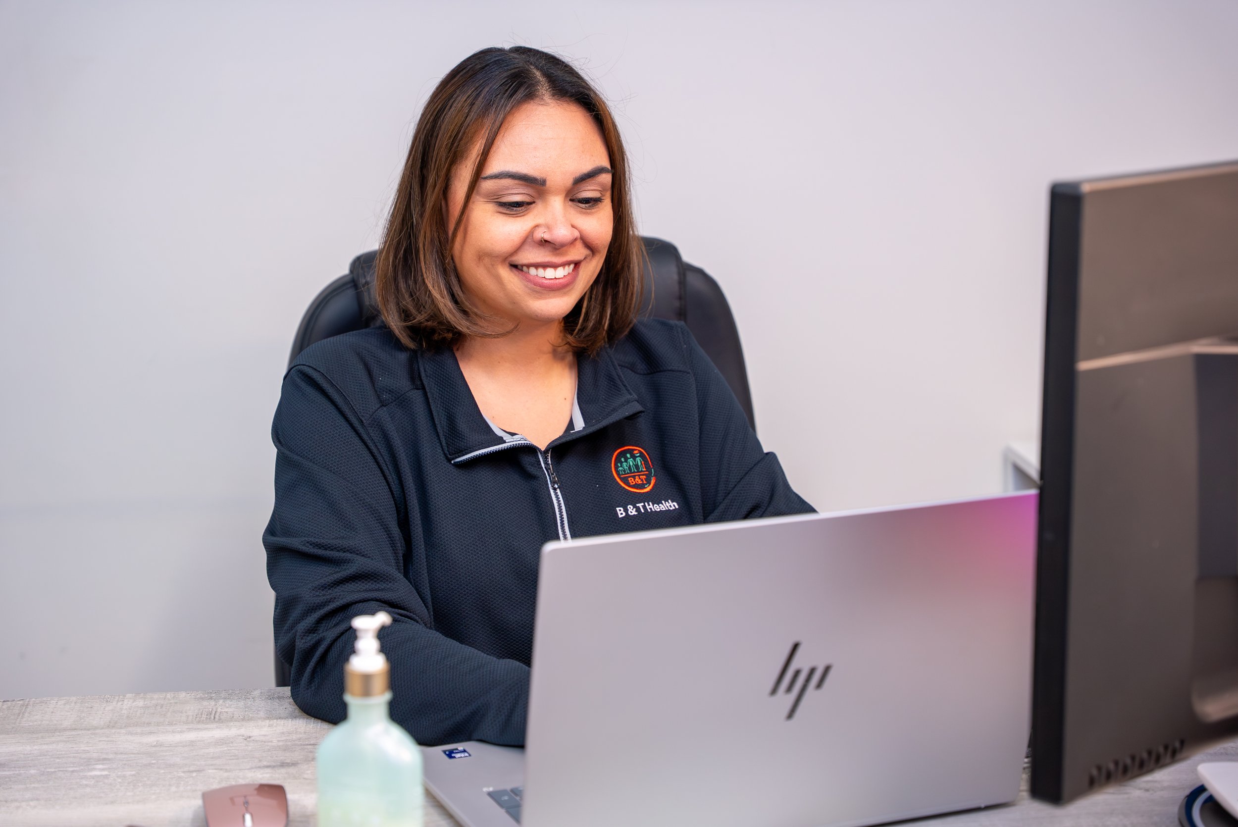 A woman with shoulder-length brown hair, smiling, sitting at a desk working on a silver HP laptop. She wears a black jacket with a logo that says 'B & T Health', there is a monitor and a hand sanitizer bottle on the desk.