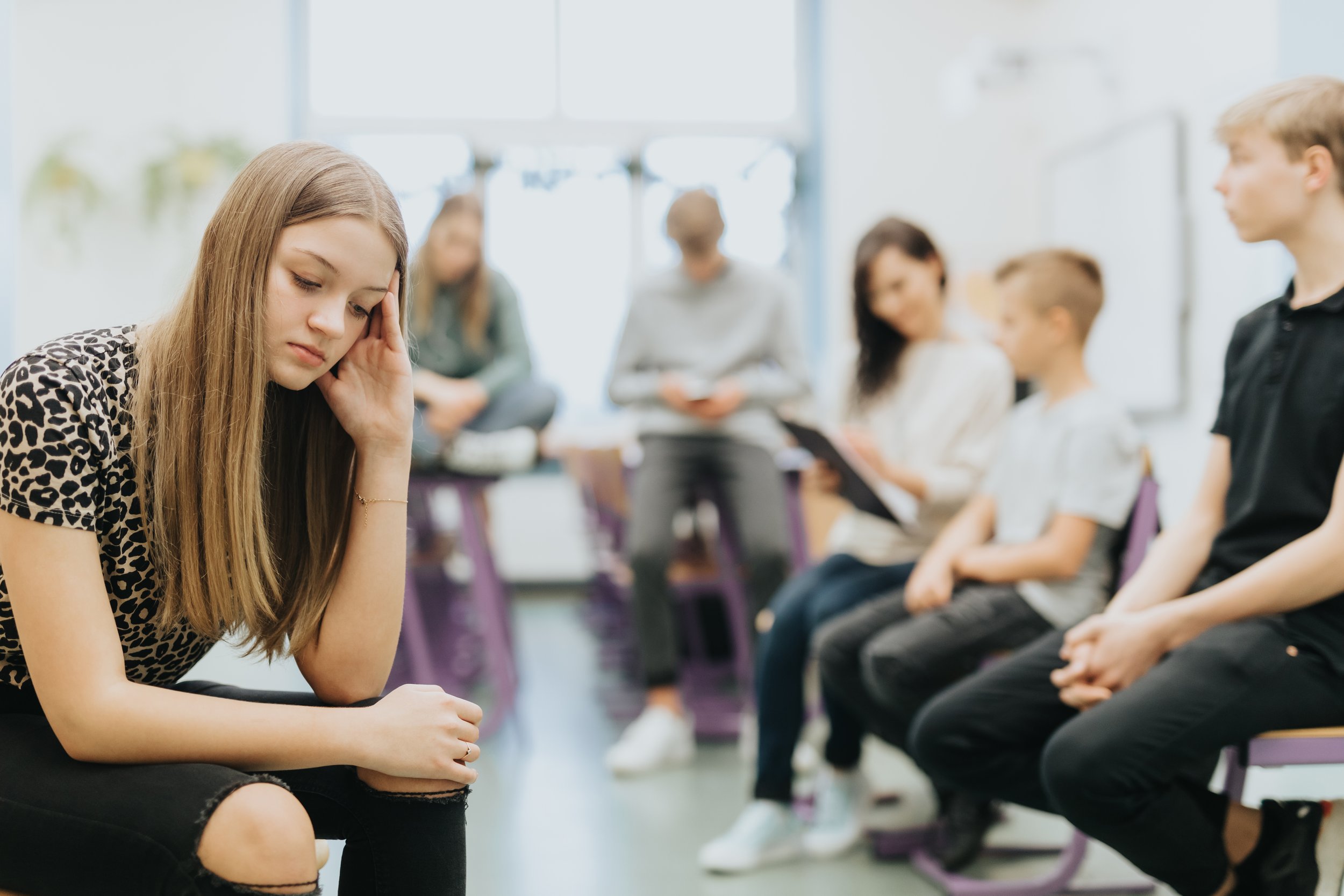 A young girl sitting with her head resting on her hand, appearing upset or distressed, in a classroom or group setting with other students seated nearby.
