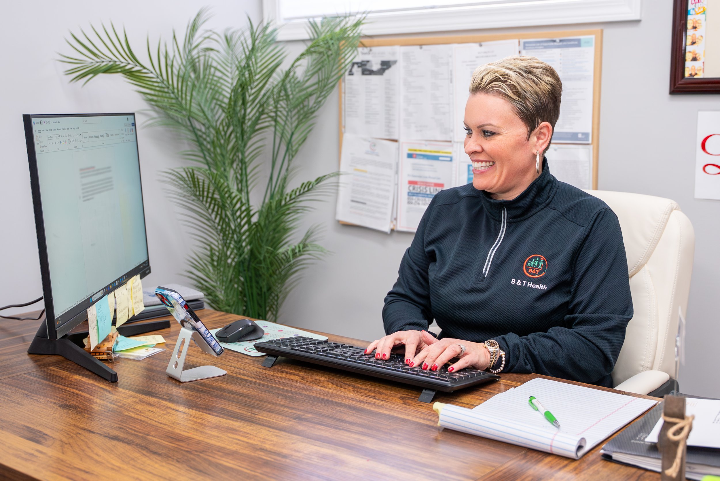 A woman with short blonde hair, wearing a black B & T Health jacket, sitting at a wooden desk and smiling while typing on a keyboard. A large desktop monitor, a smartphone on a stand, a computer mouse, and a notepad with a green pen are on the desk. There is a tall plant and a bulletin board with papers in the background.