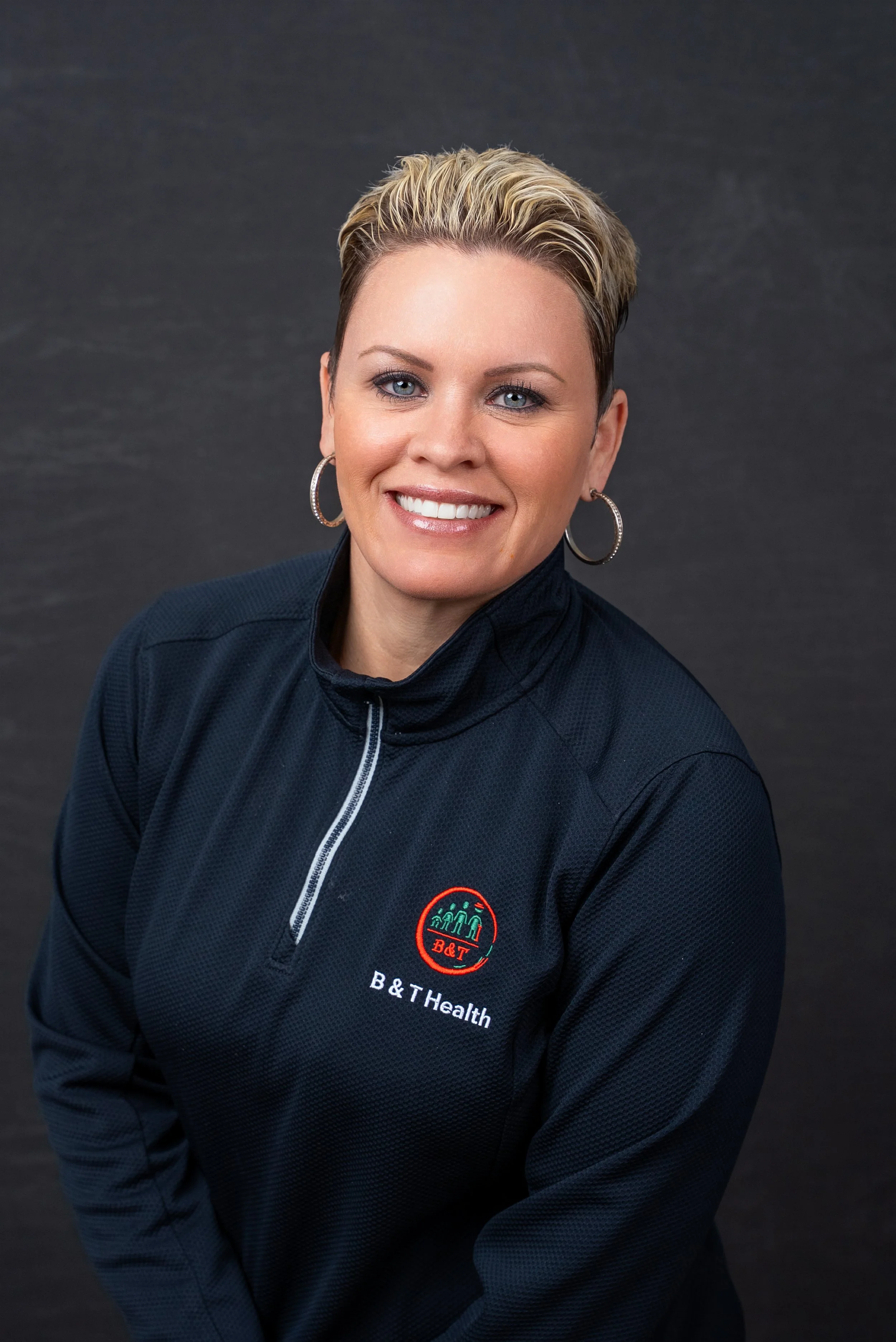 A woman with short blonde hair smiling, wearing hoop earrings and a black sports jacket with B&T Health logo, against a dark background.