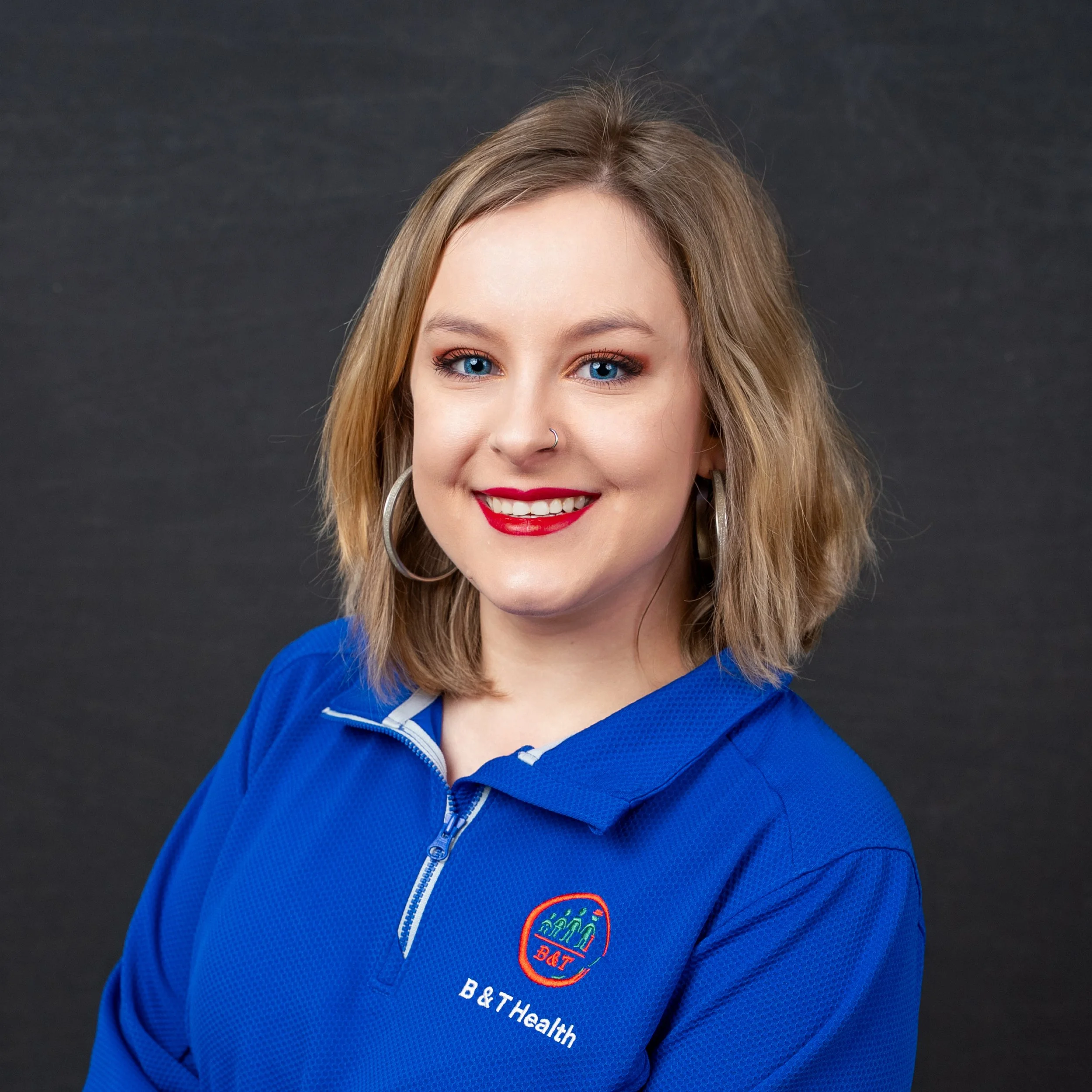 A young woman with blonde hair, blue eyes, and bright red lipstick, smiling at the camera. She is wearing a royal blue zip-up shirt with a logo that reads 'B&T Health' and large hoop earrings, standing against a dark background.