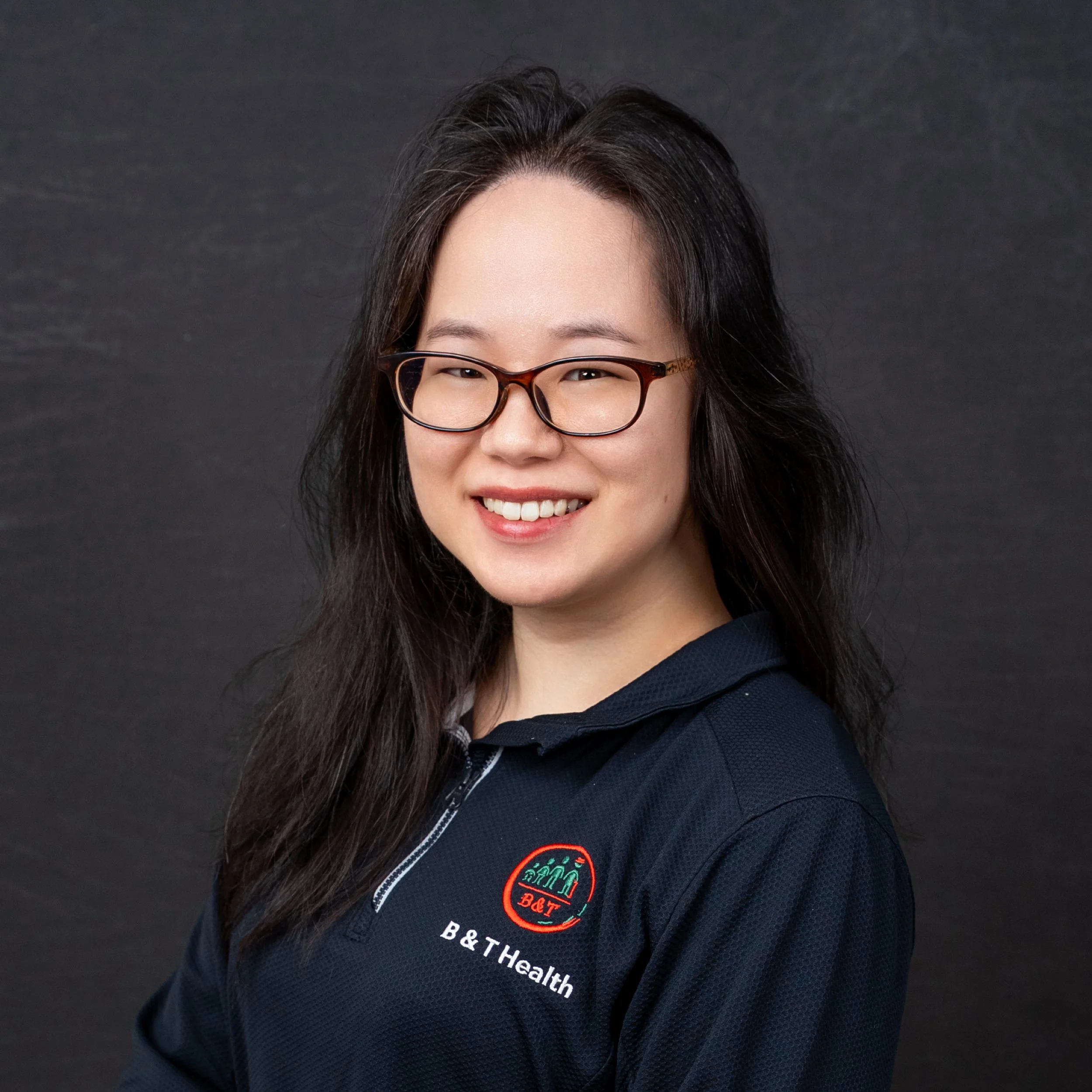 A woman wearing glasses and a navy blue polo shirt with the B&T Health logo, smiling against a dark background.