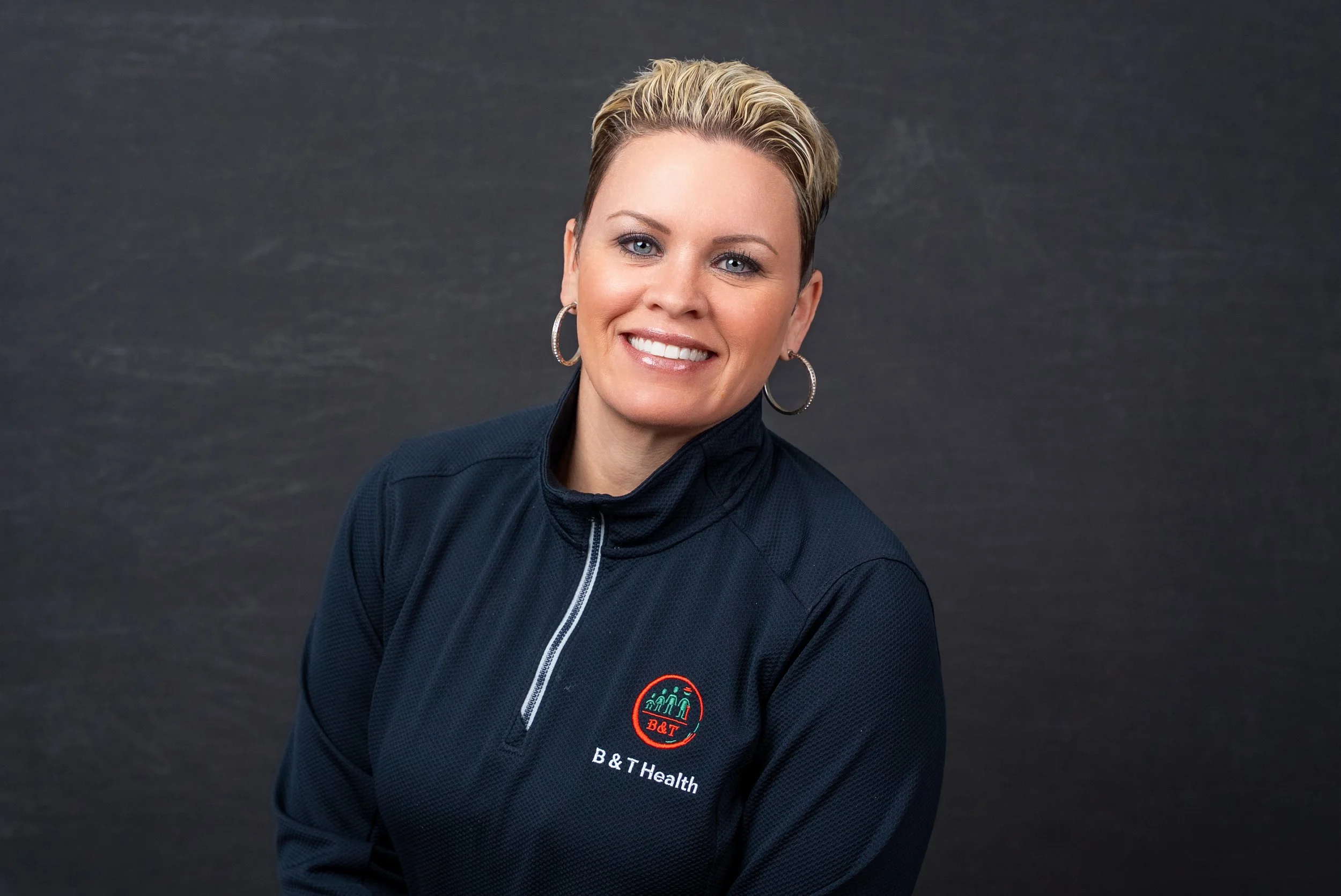 A woman with short blonde hair smiling, wearing a dark blue sports jacket with a logo that reads 'B&T Health' against a dark gray background.