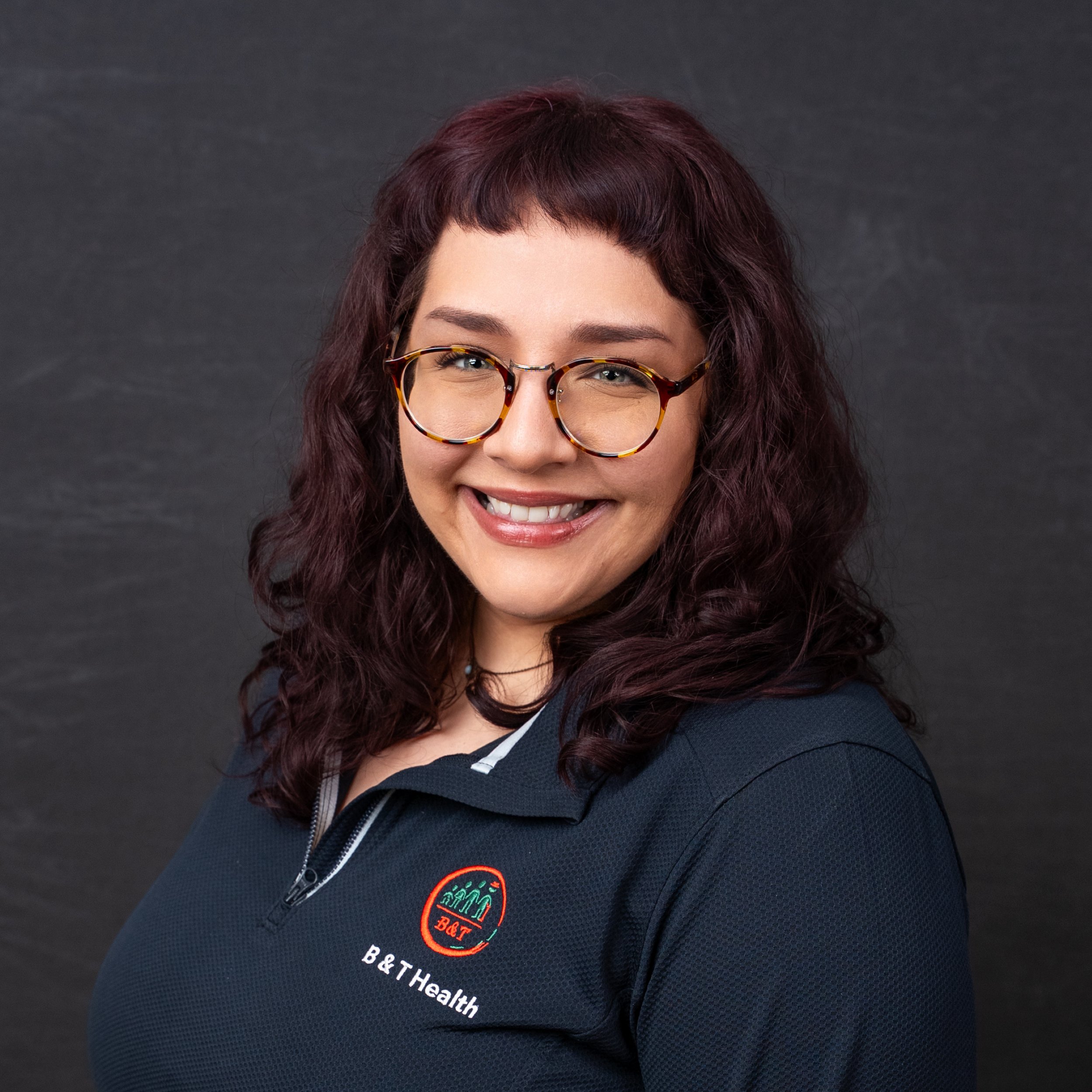 A woman with curly purple hair wearing glasses and a dark navy polo shirt with a B&T Health logo, smiling against a black background.