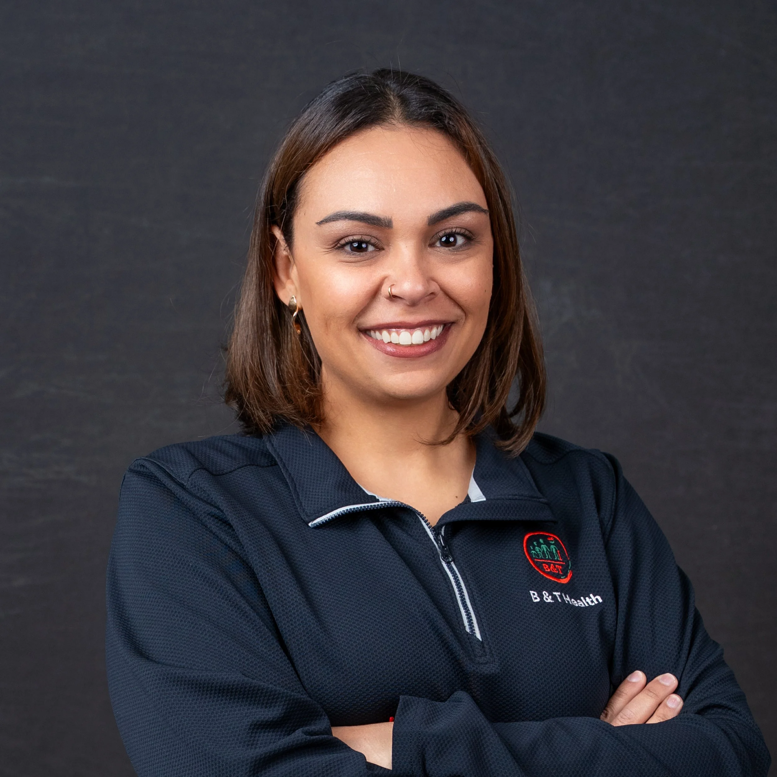 A young woman with shoulder-length brown hair smiling and crossing her arms, wearing a black athletic jacket with a logo and the text 'B & T Health' on it, standing against a dark background.