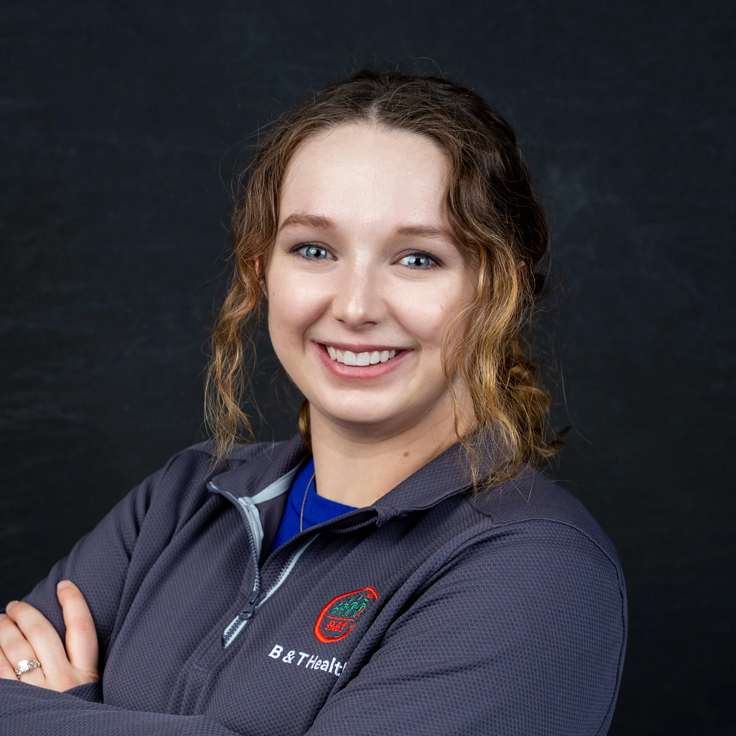 A young woman with wavy light brown hair, blue eyes, and fair skin smiling and wearing a dark gray jacket with a zipper, a blue shirt underneath, and a ring on her finger. The background is black.