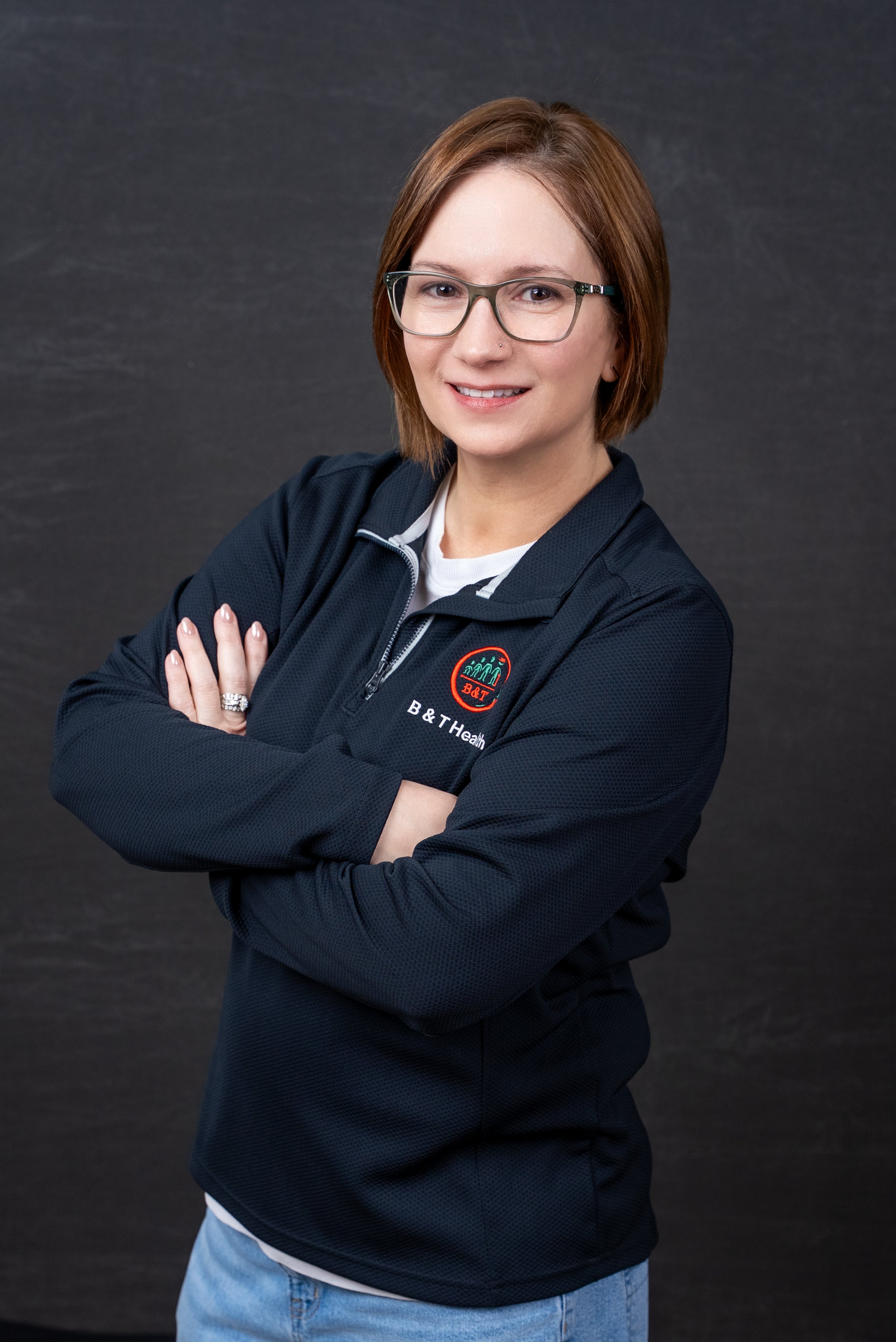 A woman with short brown hair, glasses, and a nose ring, wearing a black zip-up sports jacket with a B & T logo, standing with arms crossed against a dark background.