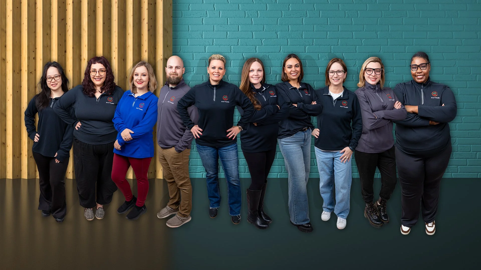 A diverse group of ten people standing in a line indoors against a blue brick wall and wooden paneling, all wearing casual and team-branded clothing, smiling, and with arms crossed or hands on hips.