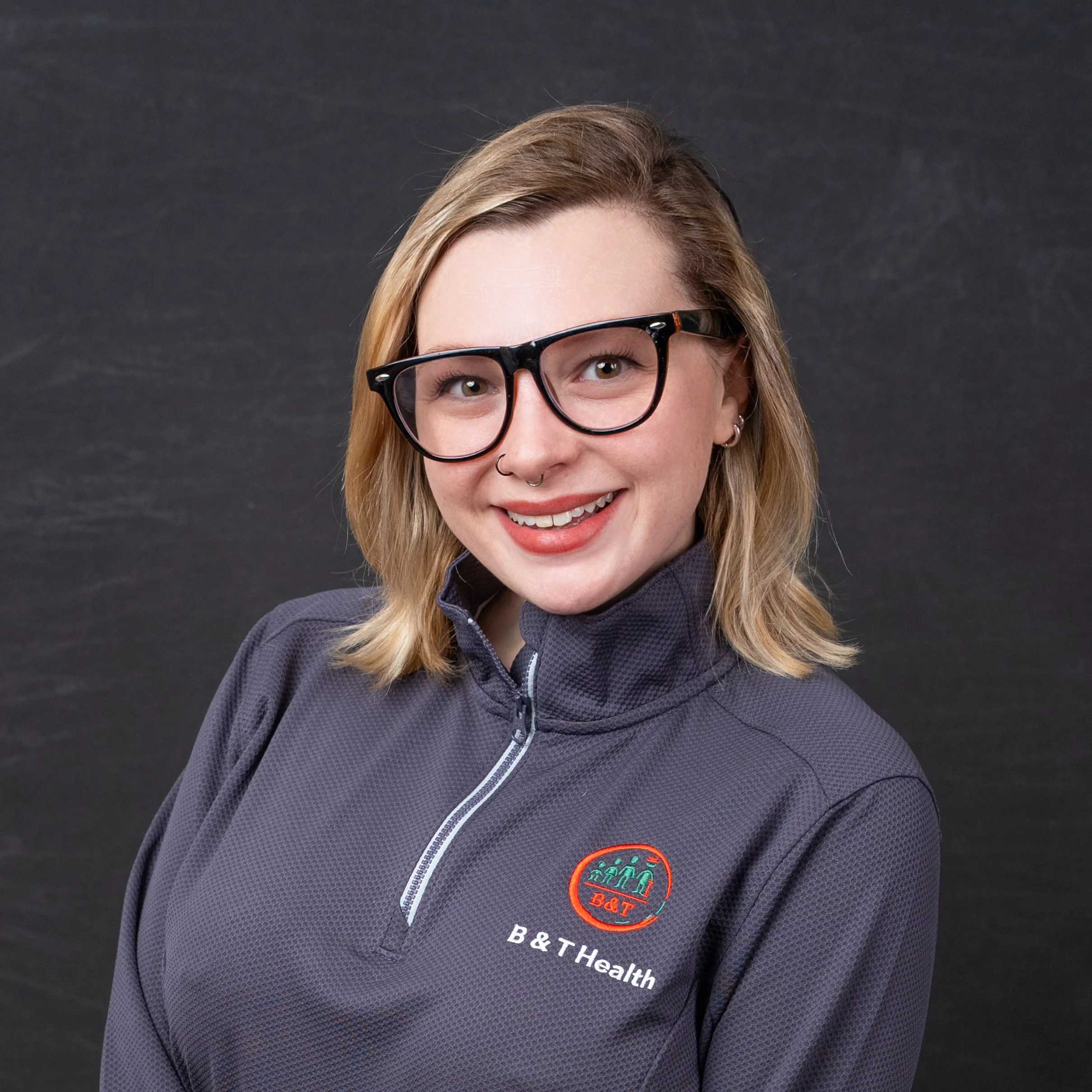 A woman with blonde hair, glasses, and a septum piercing smiling in front of a dark background, wearing a gray B&T Health jacket.