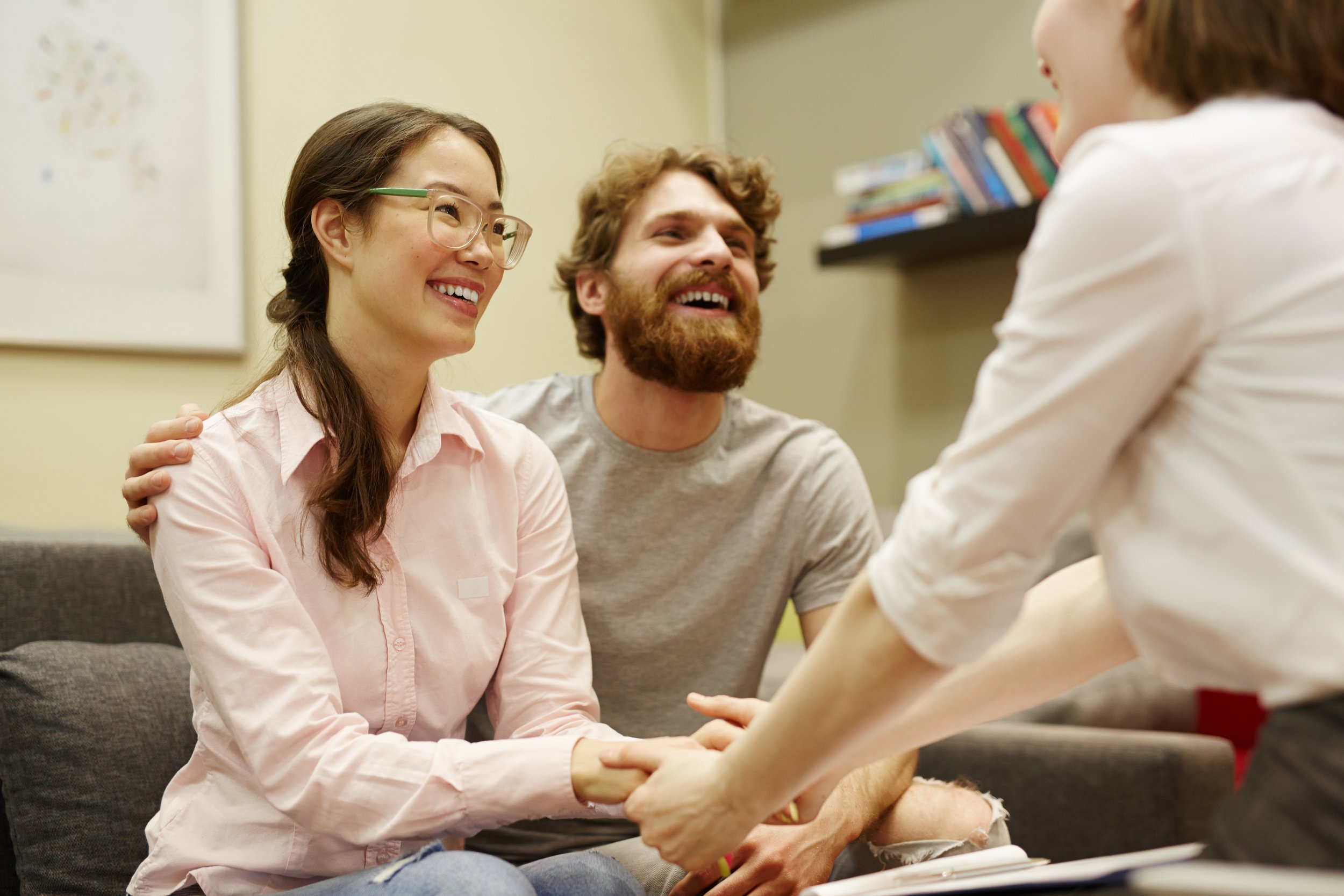 A woman with glasses and a man with a beard shaking hands with a woman across from them, smiling and engaging in a conversation.