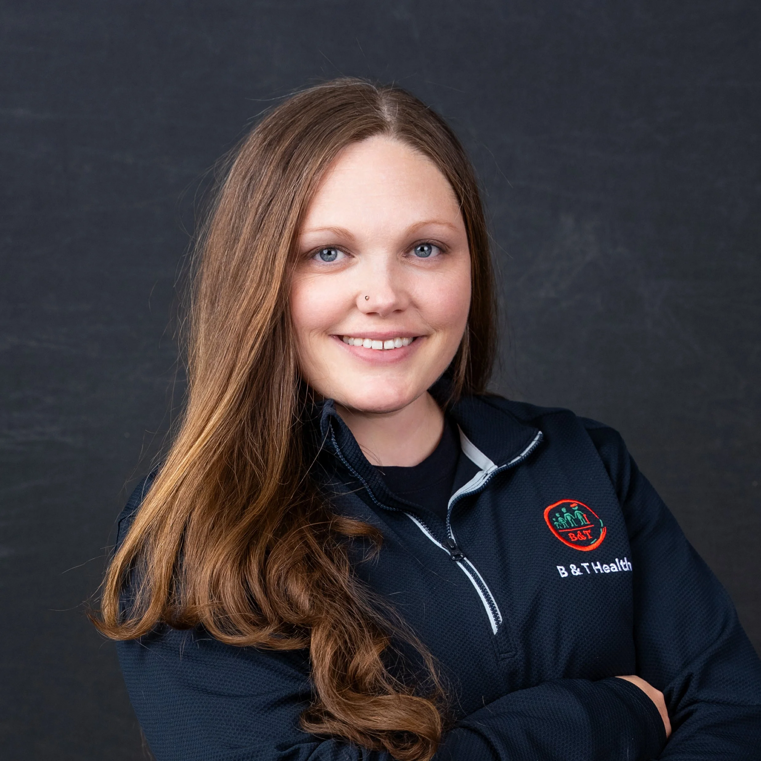 A young woman with long, wavy brown hair wearing a black zip-up jacket with a B & T Health logo, smiling with crossed arms against a dark background.
