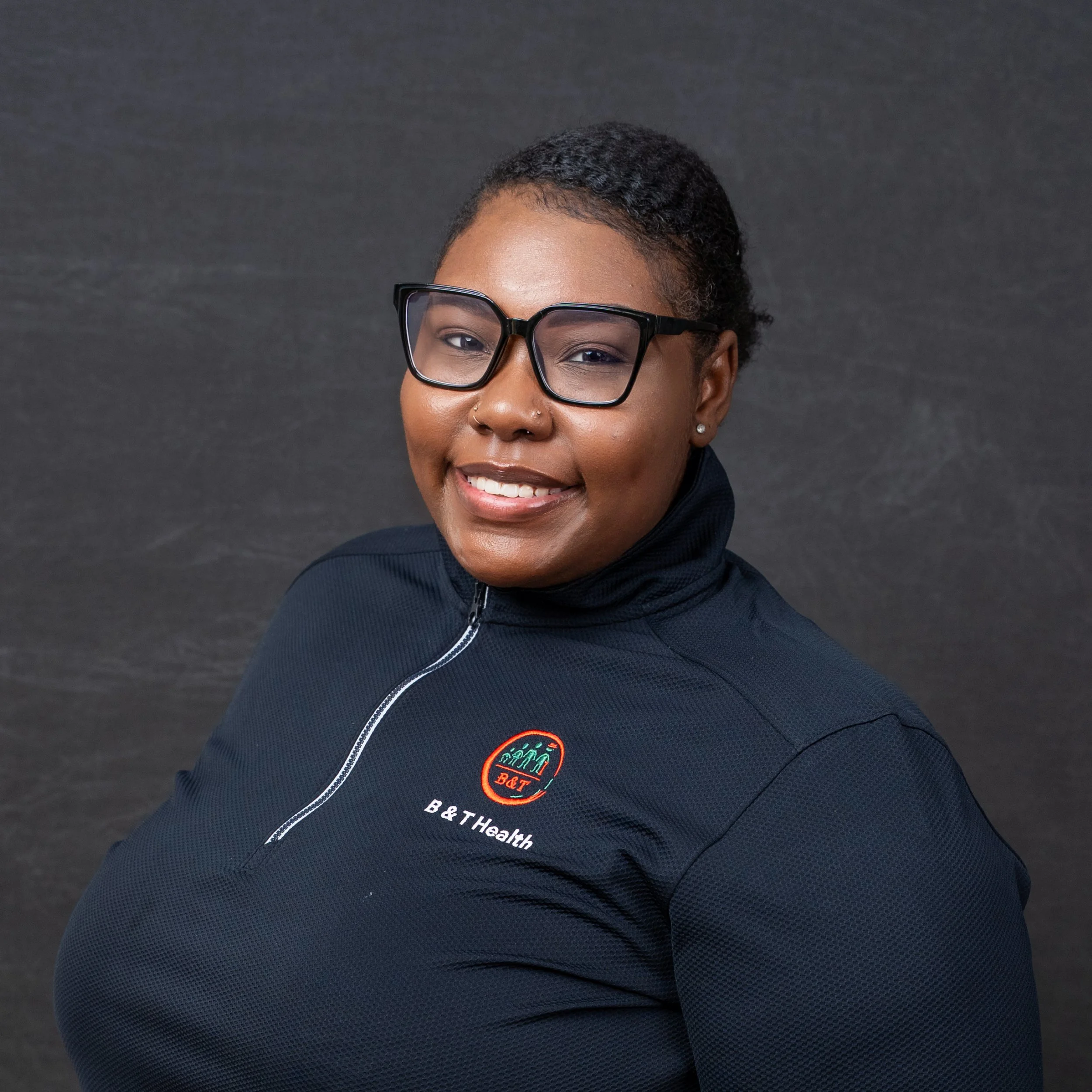 A woman with short black hair, glasses, and a nose piercing smiling, wearing a navy sports jacket with a "B&T Health" logo, standing against a dark background.