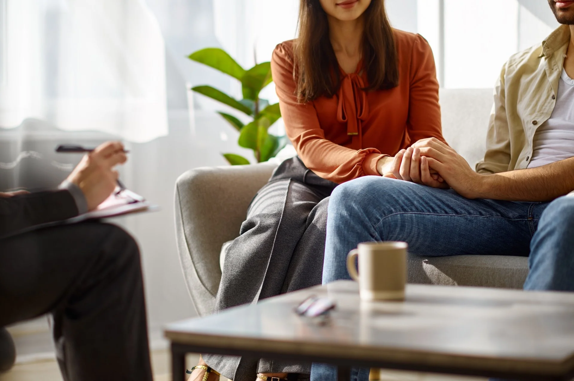 Therapist taking notes during a counseling session with a woman holding the man's hand in a cozy, well-lit room with plants and coffee mugs.