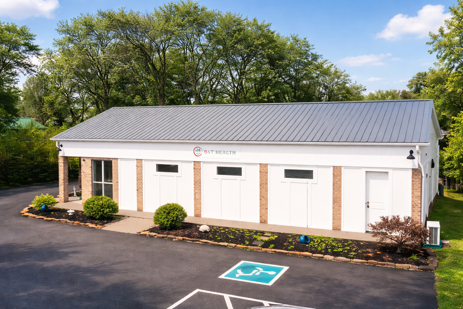 A white building with brick accents and a metal roof, with a sign reading "B&T Health" on the front, surrounded by trees and a landscaped area with shrubs and decorative rocks. There is a parking space for handicapped in the foreground.