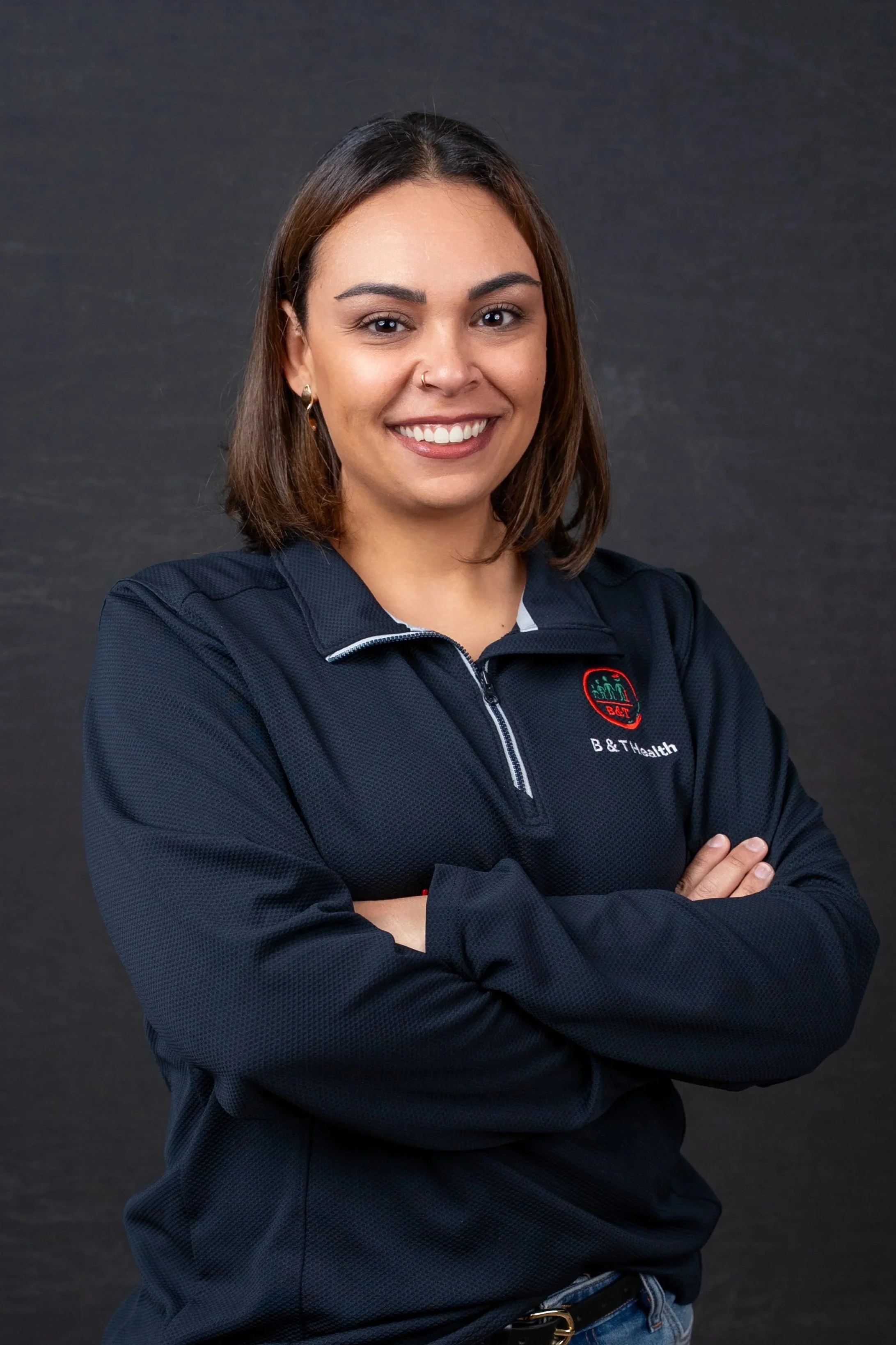 A young woman with shoulder-length brown hair, smiling, wearing a navy blue quarter-zip jacket with a logo and the words "B & T Health," and standing with arms crossed against a dark background.