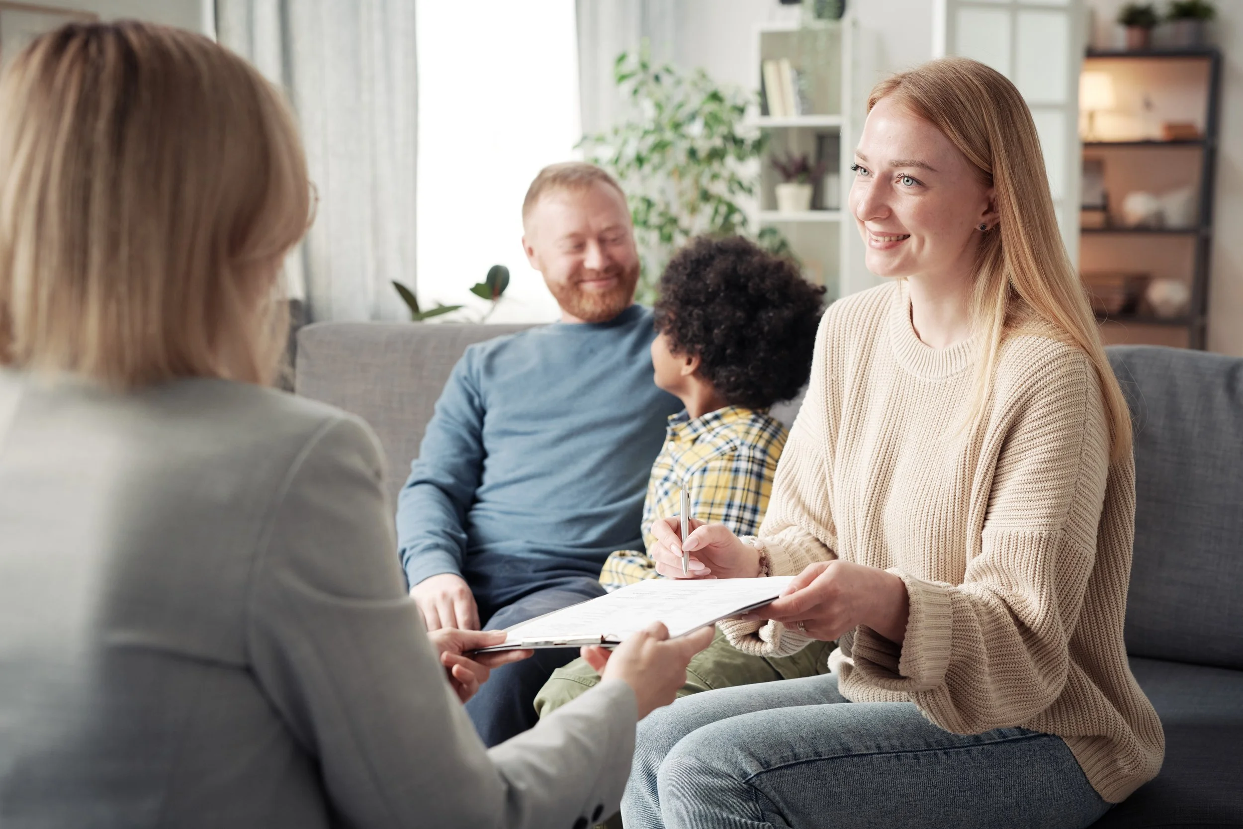 A woman in a beige sweater taking notes while talking to a young girl with curly hair, sitting on a gray sofa. In the background, a man with a beard and a boy with curly hair sit on the sofa, smiling. The room is bright and decorated with plants and shelves.