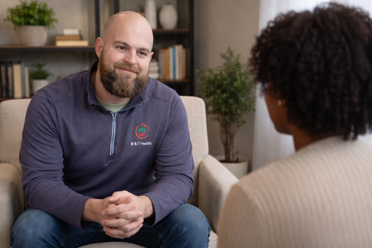 A man with a beard and shaved head is sitting on a beige couch, talking to a woman with curly hair. He is wearing a purple fleece with a logo and the words "B & T Health." The background features a bookshelf with books and potted plants.