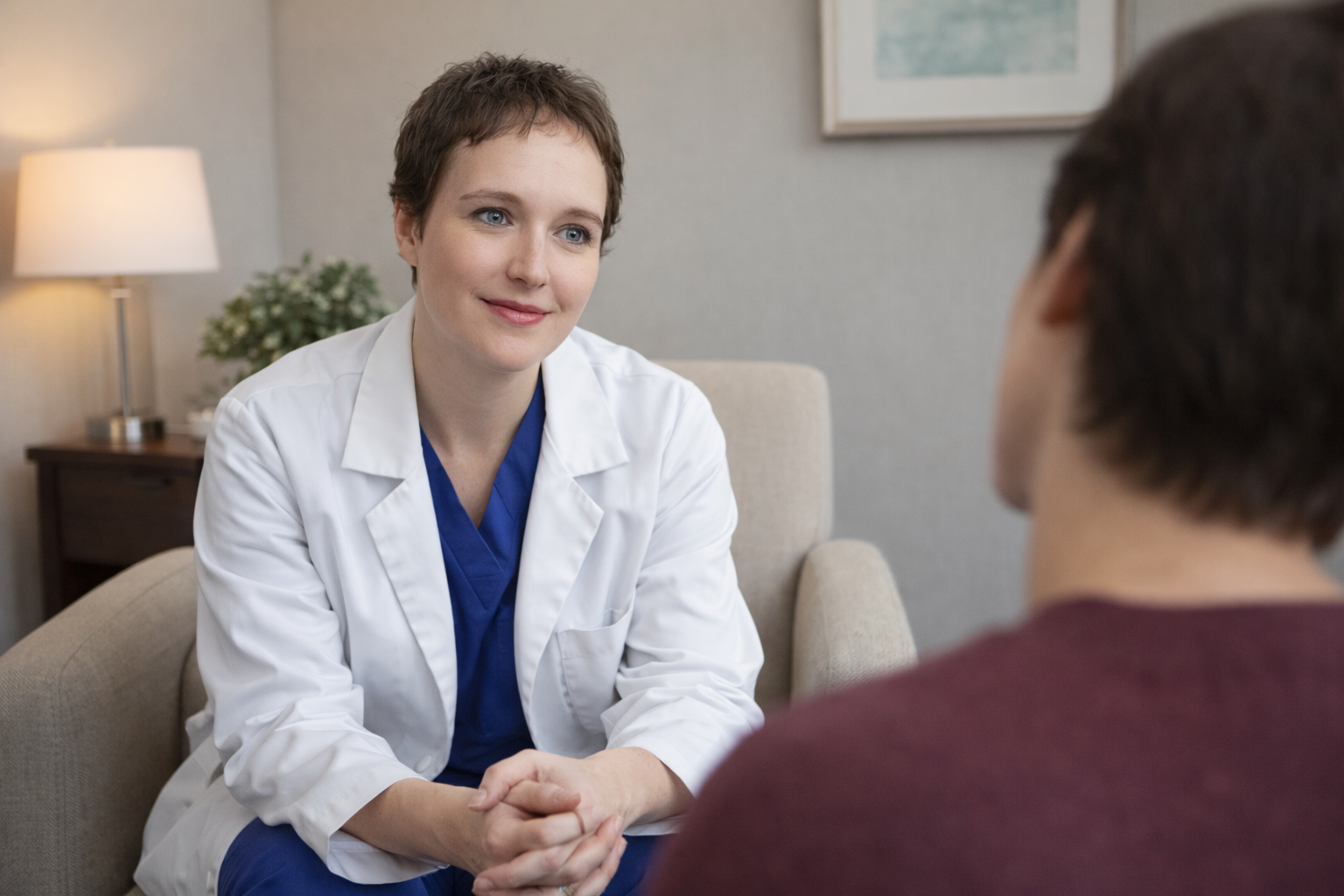 A female doctor with short brown hair and blue eyes wearing a white coat and blue scrubs, sitting on a beige armchair and talking to a male patient in a medical consultation room.