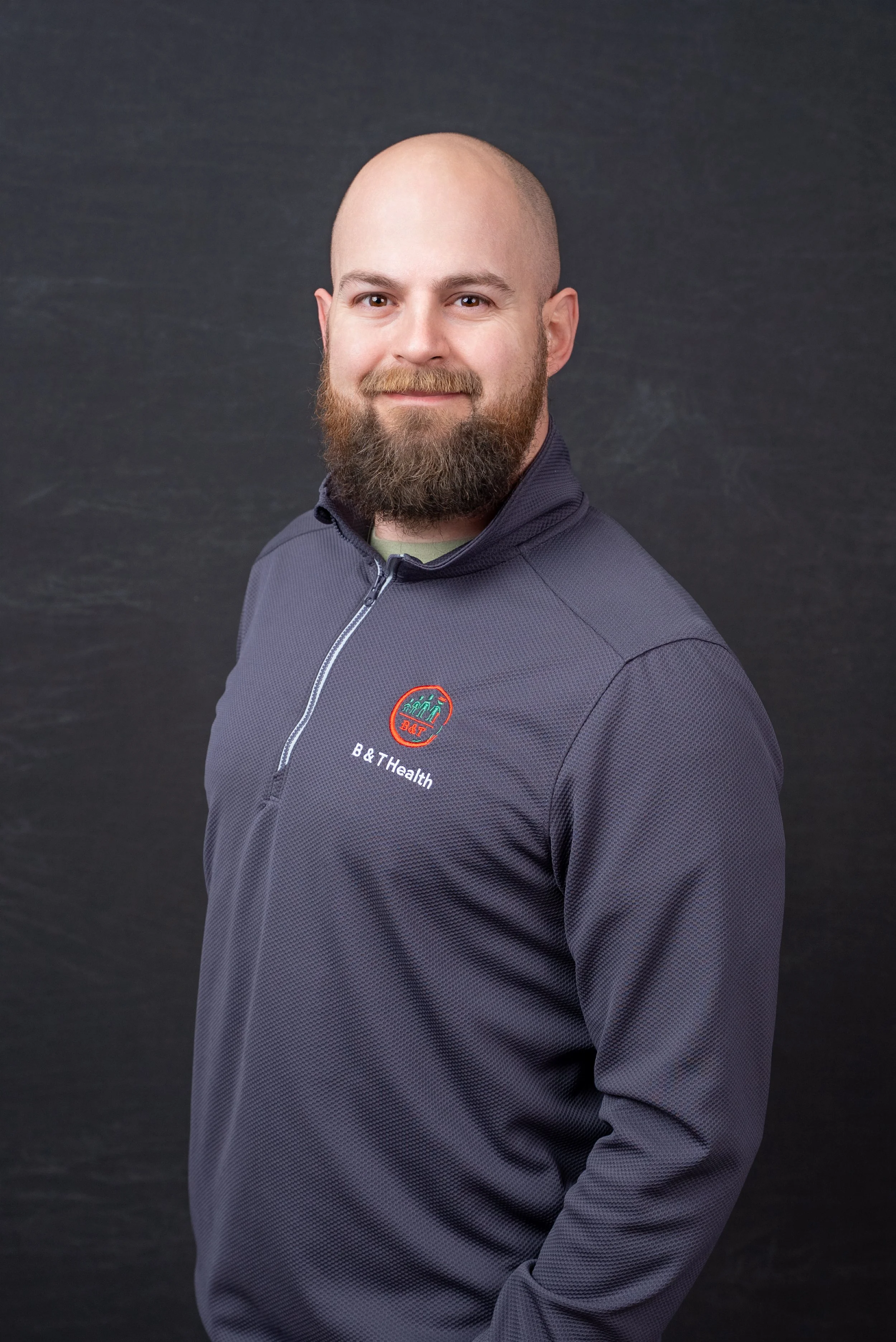 A man with a brown beard and a shaved head, wearing a dark gray zip-up jacket with a 'B&T Health' logo, standing against a dark background.