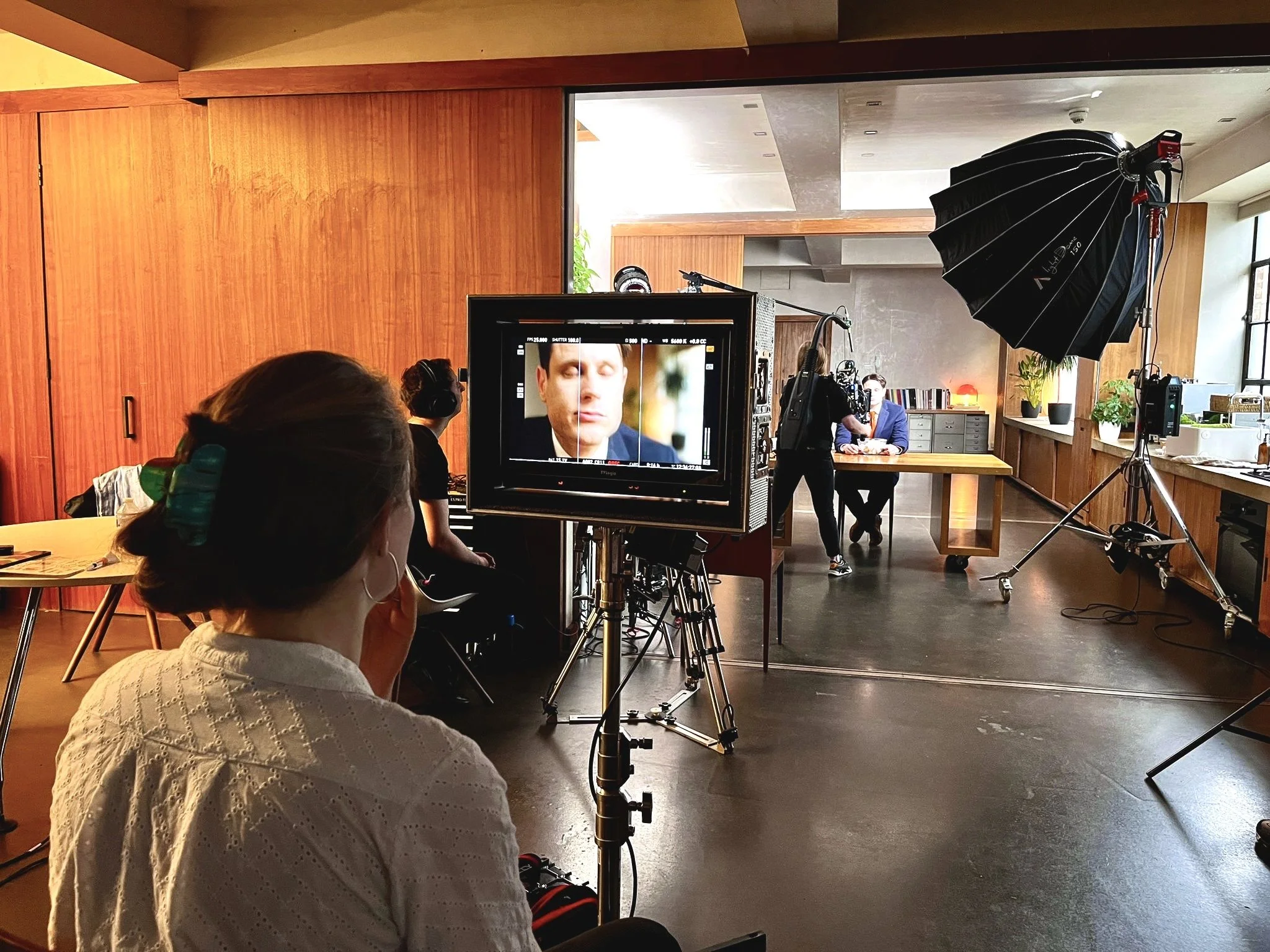 Film crew filming a scene in a studio with a camera, monitor, and lighting equipment. A woman with her hair in a bun and a hair clip is seated in front of the camera, and a man and woman are sitting at a table in the background.