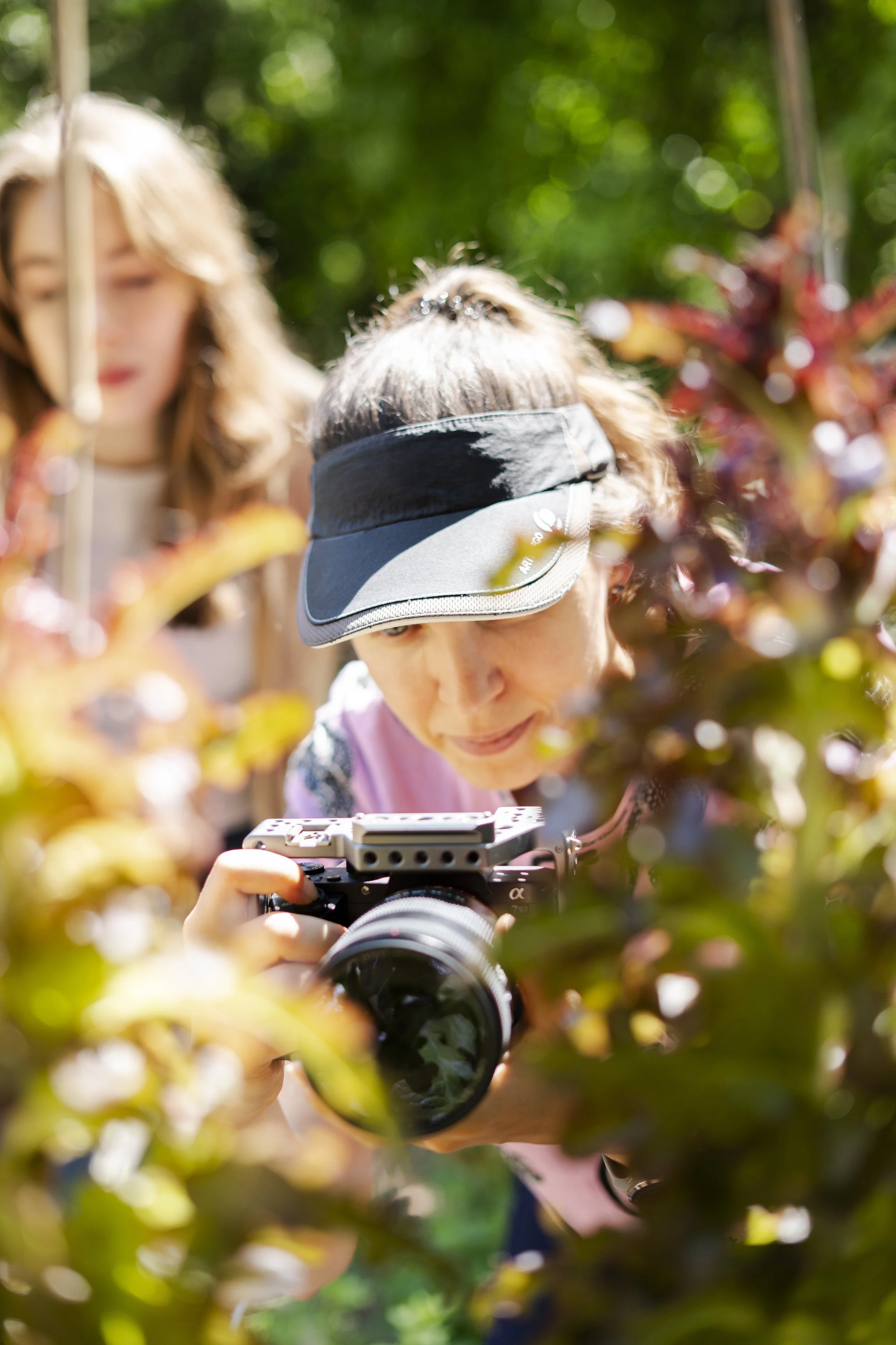 A woman taking a photograph with a camera outdoors, surrounded by bushes, with two other women in the background, in a sunny setting.