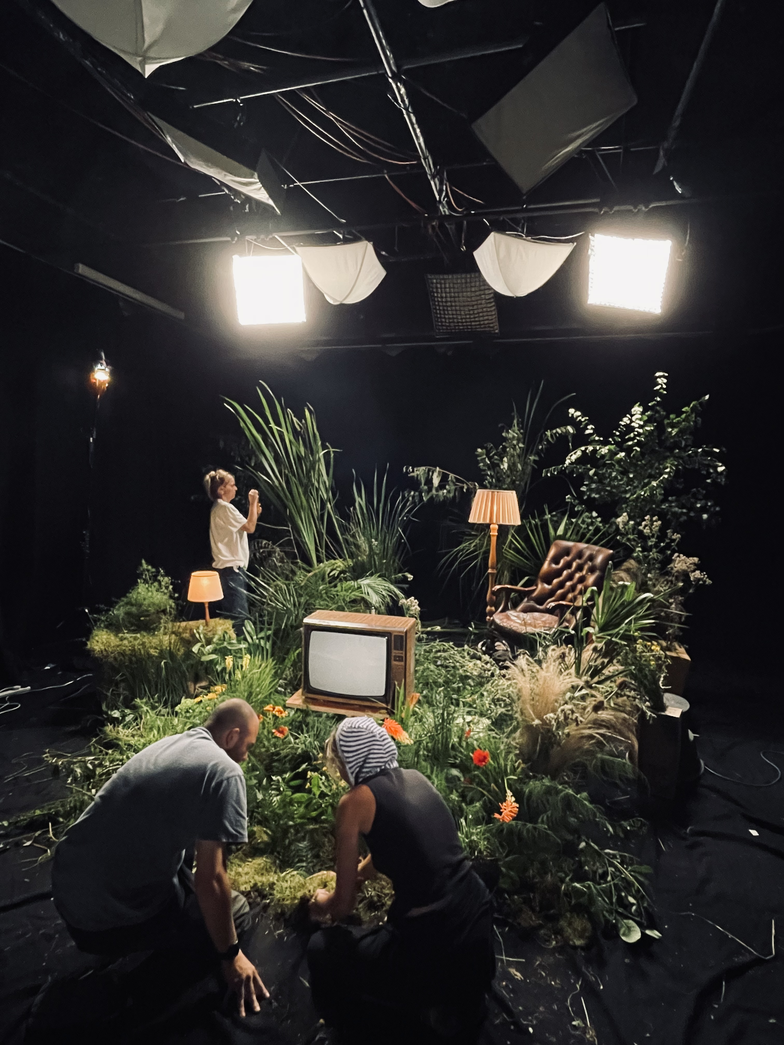 People setting up a stage surrounded by plants and vintage furniture, with studio lights overhead.