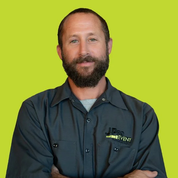 Man smiling and posing with arms crossed, wearing a dark grey shirt with a logo that says 'JPRO EVENT MANAGEMENT' on a green background.