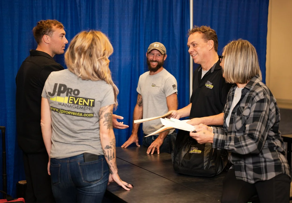 Group of five people converse at a signing table at an event, with a woman and man holding papers and smiling.