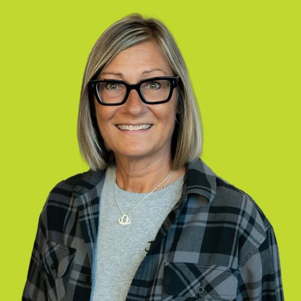 A woman wearing a plaid shirt, gray t-shirt, and a silver necklace, smiling against a lime green background.