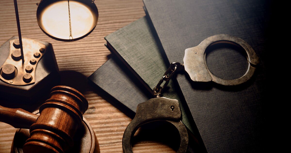 A wooden gavel, a pair of handcuffs, a desk lamp, an old rotary phones, files or black books on a wooden desk.