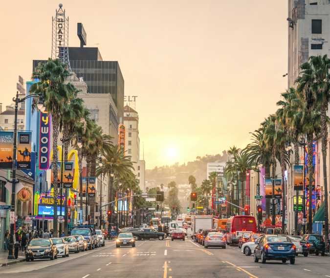 Sunset over a busy street in Hollywood, Los Angeles, with palm trees, colorful signs, and buildings lining the street.