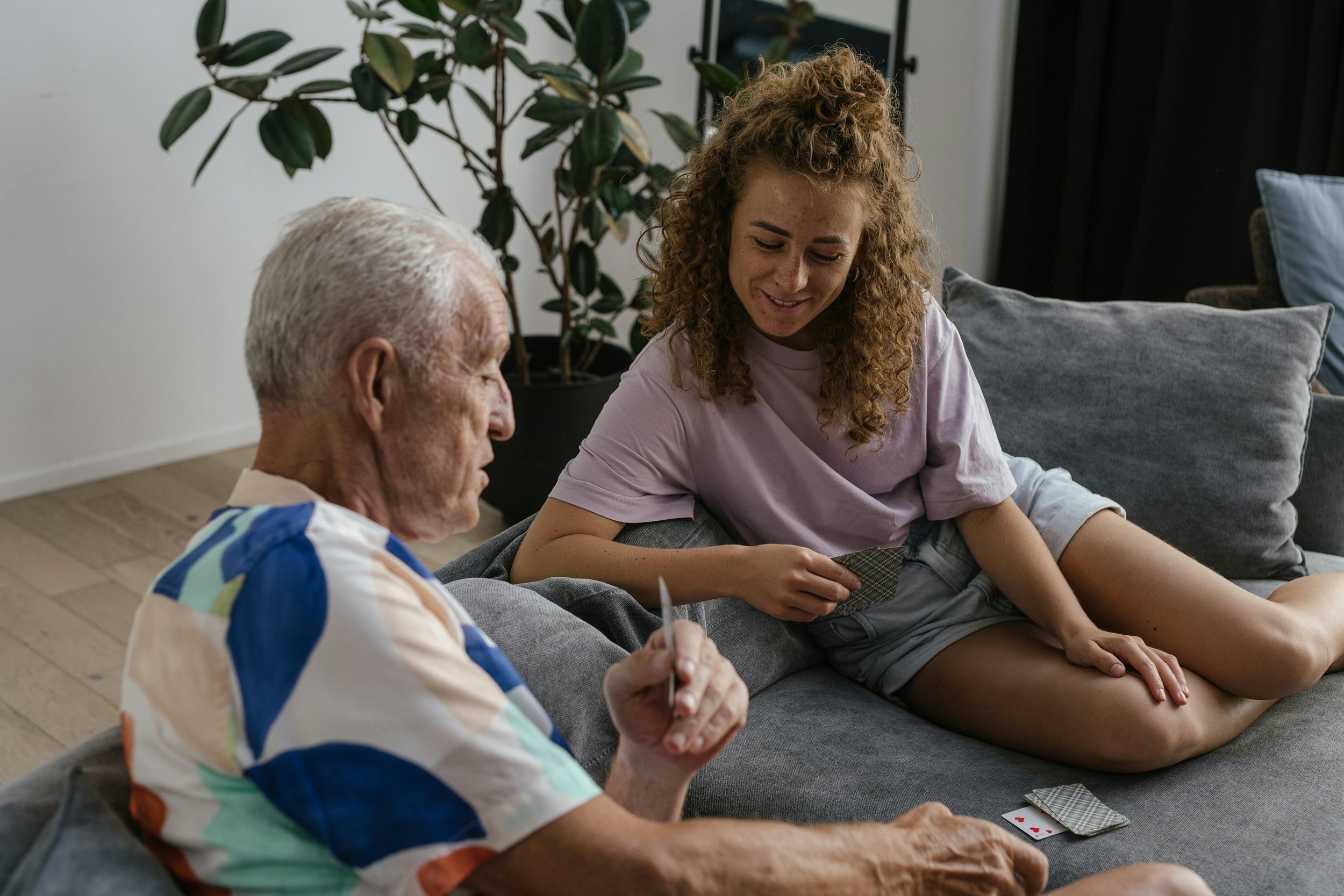 A young woman and an elderly man playing cards together on a gray sofa in a living room with a plant in the background.