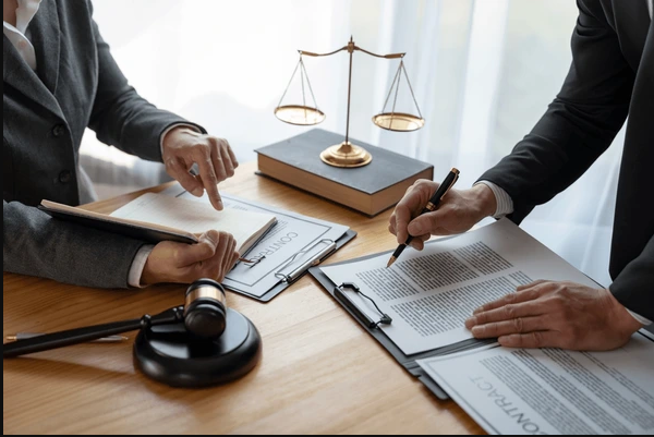 Two people in business attire reviewing legal documents at a desk with a gavel, legal scales, and a book.