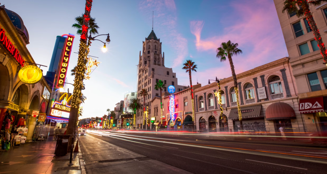 Street scene at sunset on Hollywood Boulevard, with neon signs, palm trees, and a tall building with a spire, decorated for the holidays.