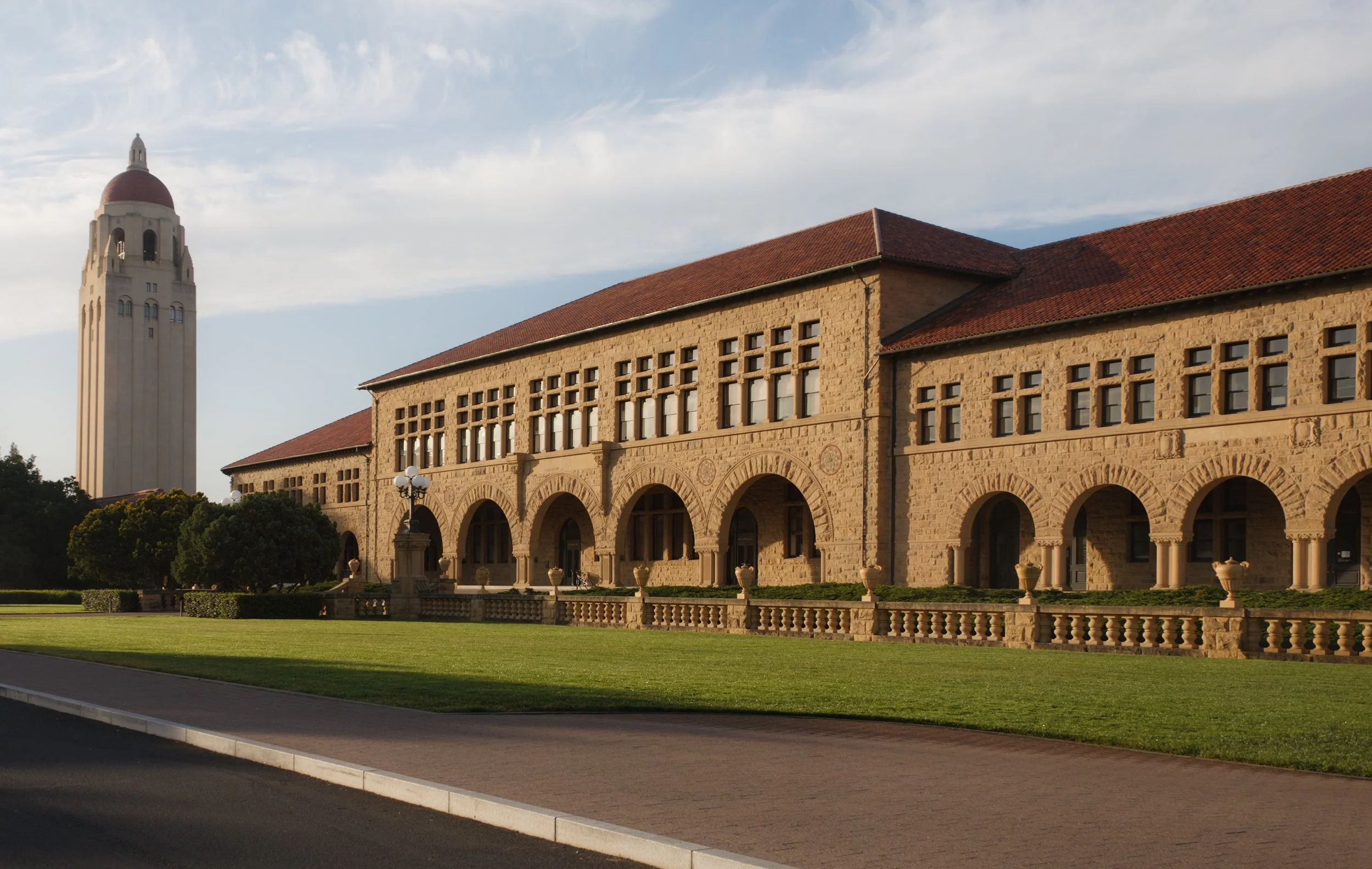 A historic stone building with arches and a red-tiled roof, alongside a tall clock tower, set against a partly cloudy sky.