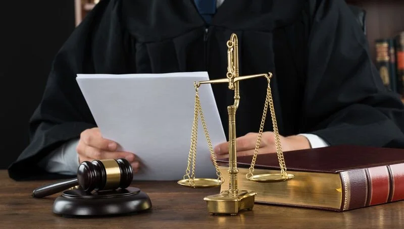 Person wearing a black robe seated at a table with legal items including a gavel, scales of justice, a thick book, and reading documents.