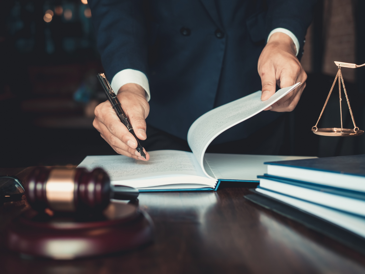 A person in a dark suit is signing a book with a black pen. There is a gavel and a stack of large books on a wooden table, and a small set of scales in the background.