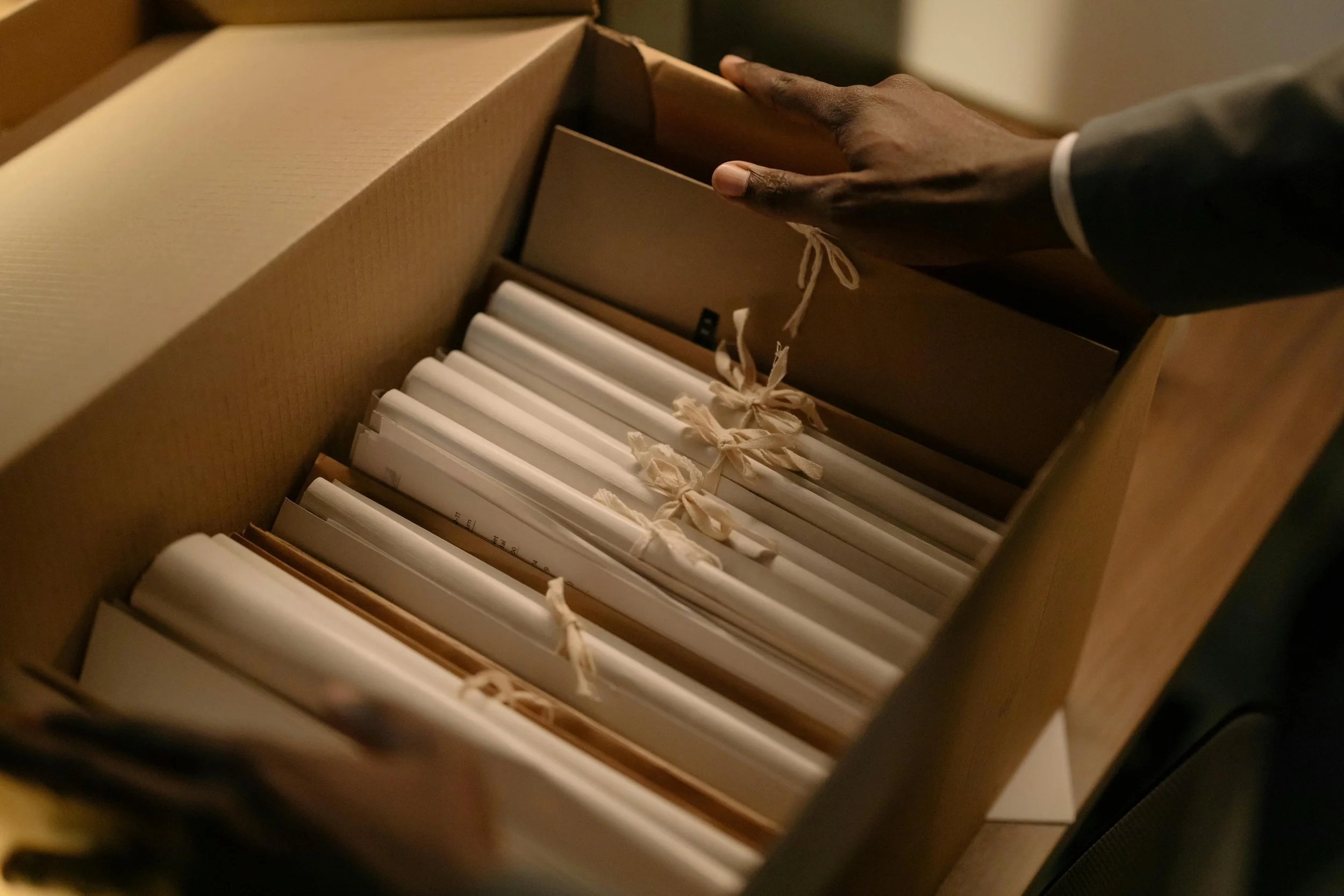 A person's hand reaching into a cardboard box filled with neatly organized file folders tied with ribbons.