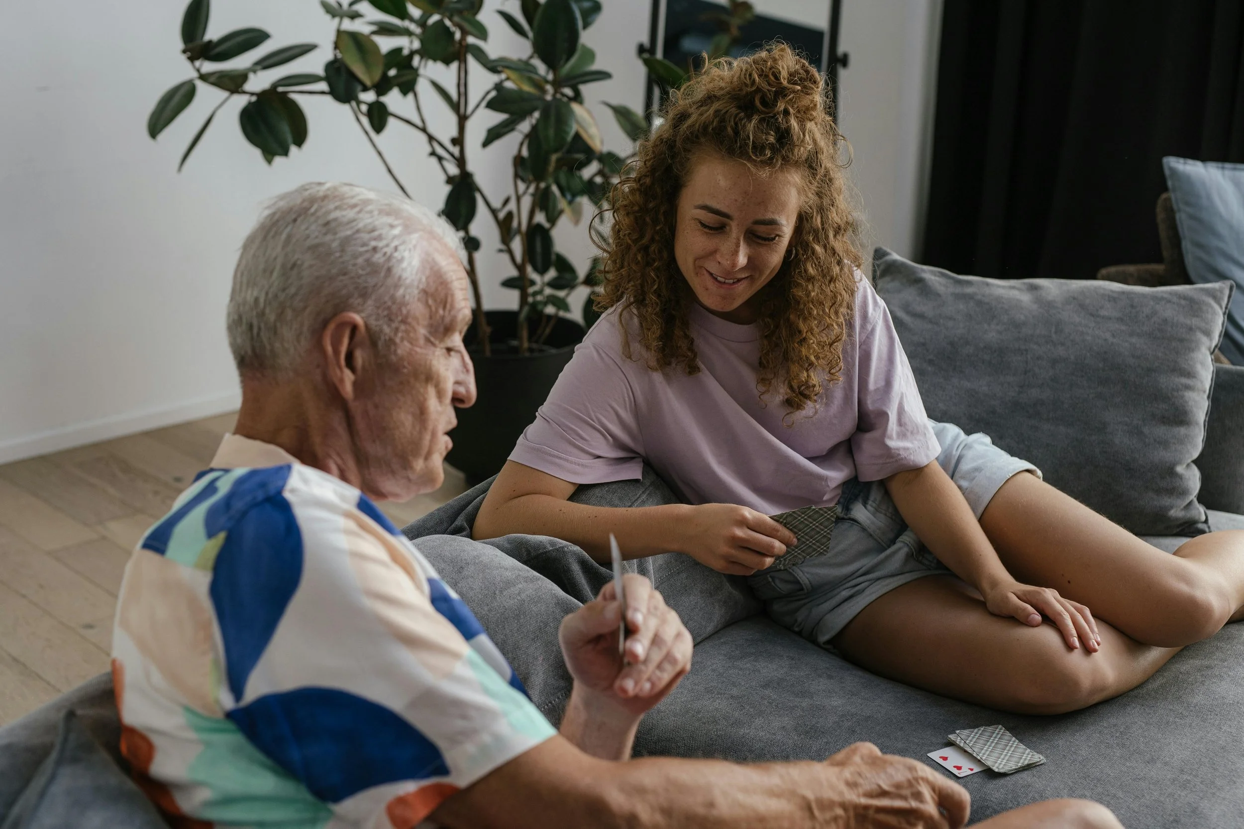 A young woman and an elderly man playing a card game on a gray couch in a living room with a plant in the background.