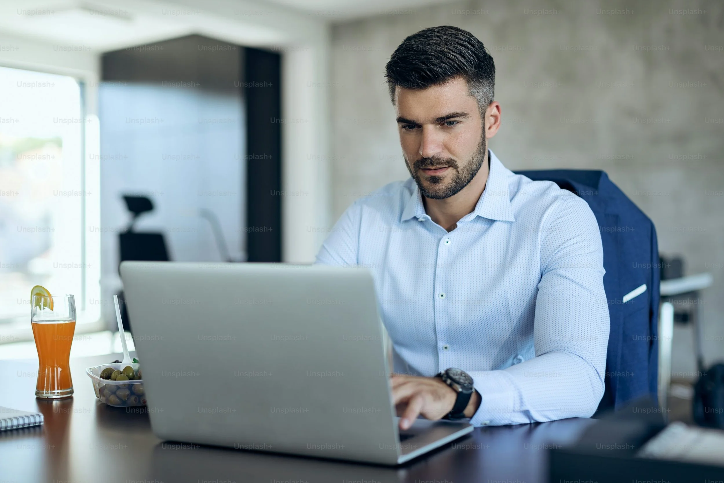 A man works on a laptop at a desk in an office, with a glass of orange juice and a bowl of olives nearby.