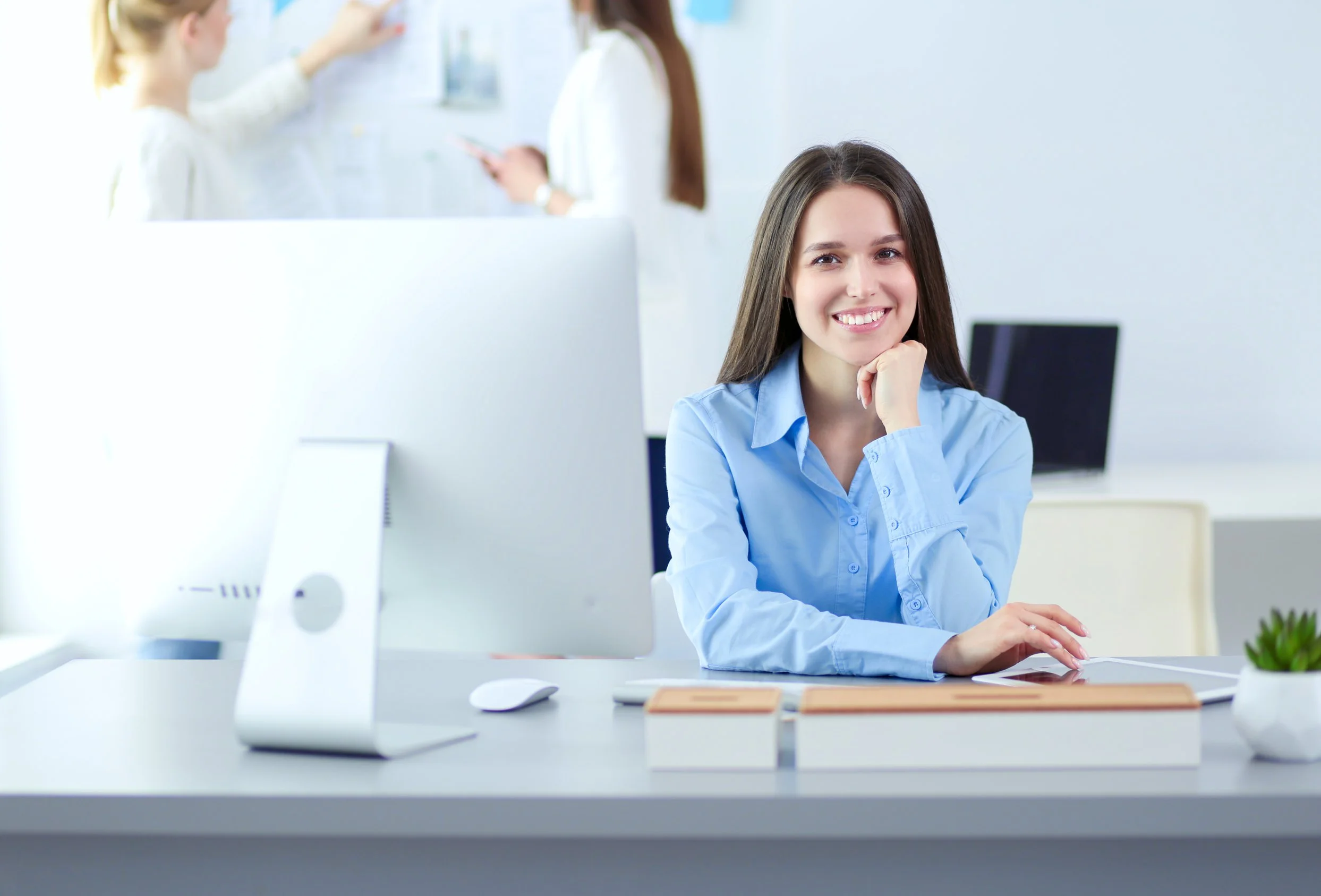 A young woman sitting at a desk in an office, smiling with her chin resting on her hand, with a computer monitor in front of her and two closed books on the desk. There are two other women in the background talking and looking at a bulletin board.