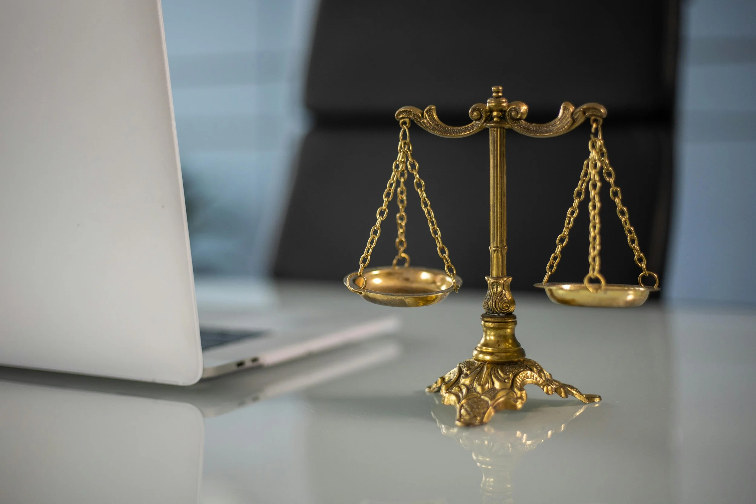 A decorative gold scale of justice placed on a white desk with a laptop nearby and a black chair in the background.