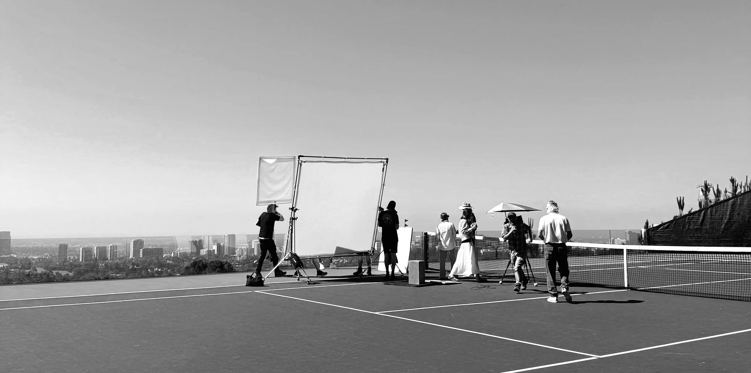 Photography crew filming a scene on an outdoor tennis court with city skyline in the background.
