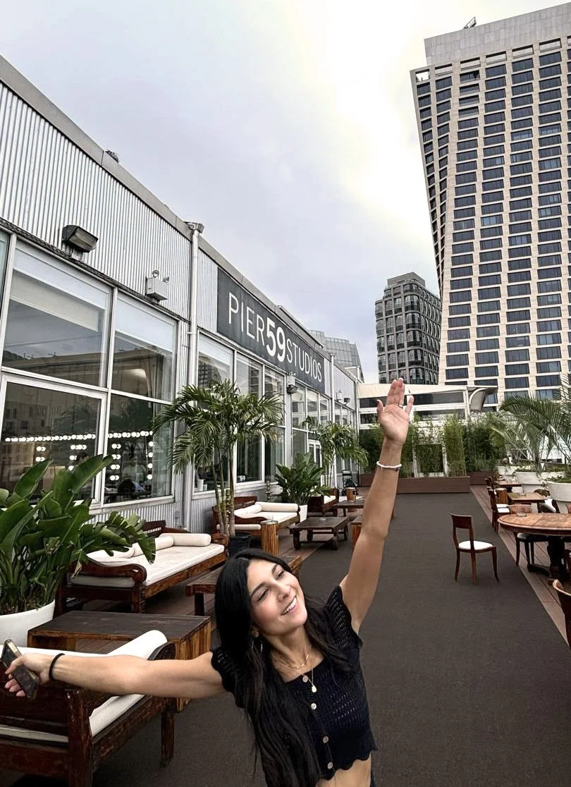 A woman smiling and stretching her arms on a rooftop patio with outdoor furniture and plants, with tall buildings and a cloudy sky in the background.