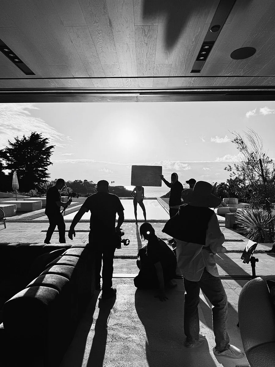 Silhouettes of a film crew on a set with a model posing outdoors, with the sun in the background, near a pool and trees.