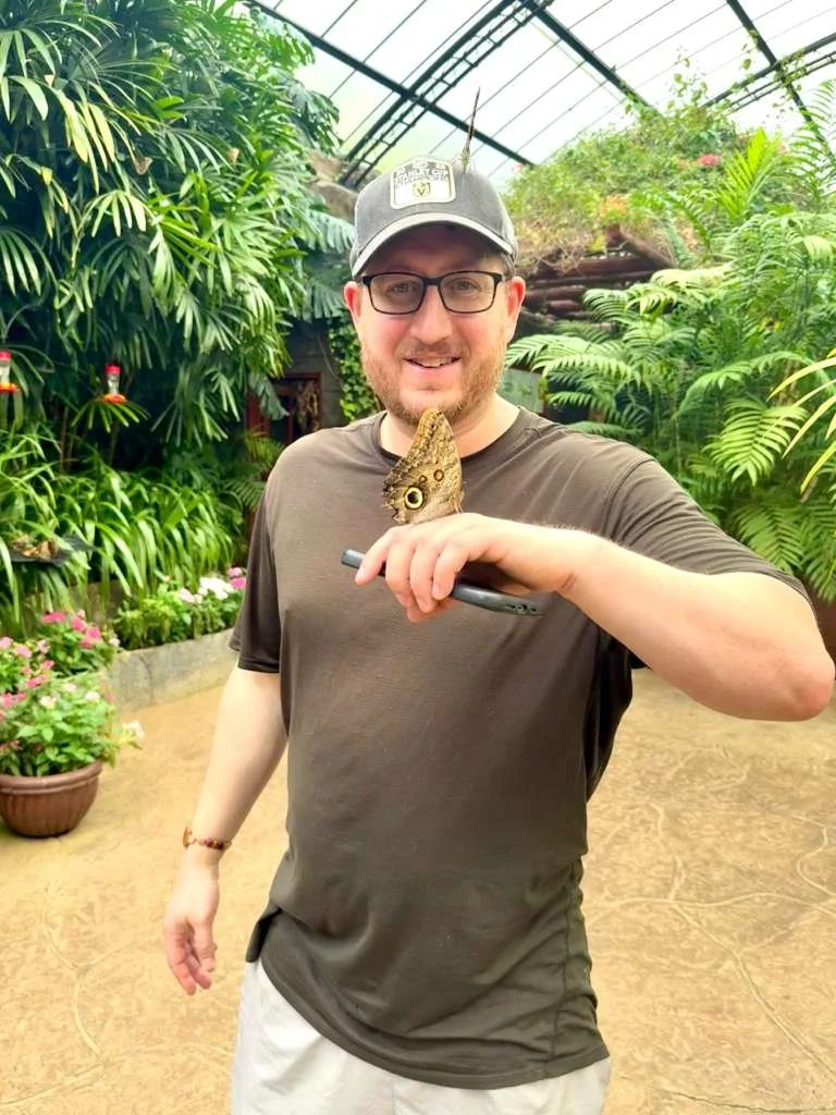 A man smiling with a large butterfly on his chest in a lush greenhouse.