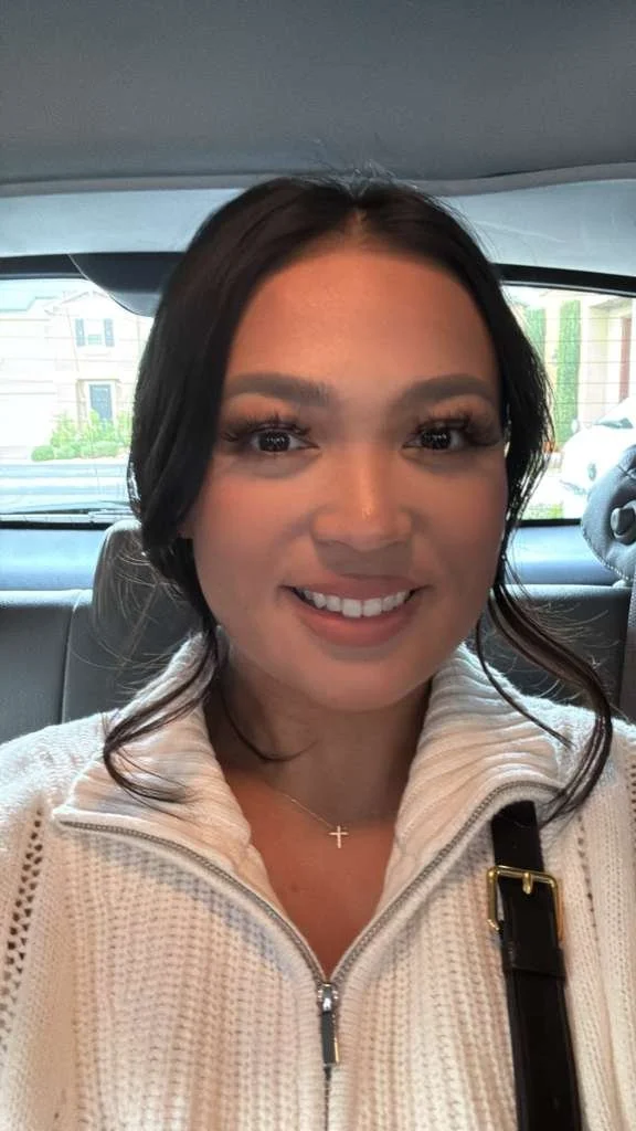 A smiling woman with dark hair in a white sweater, sitting in a car.